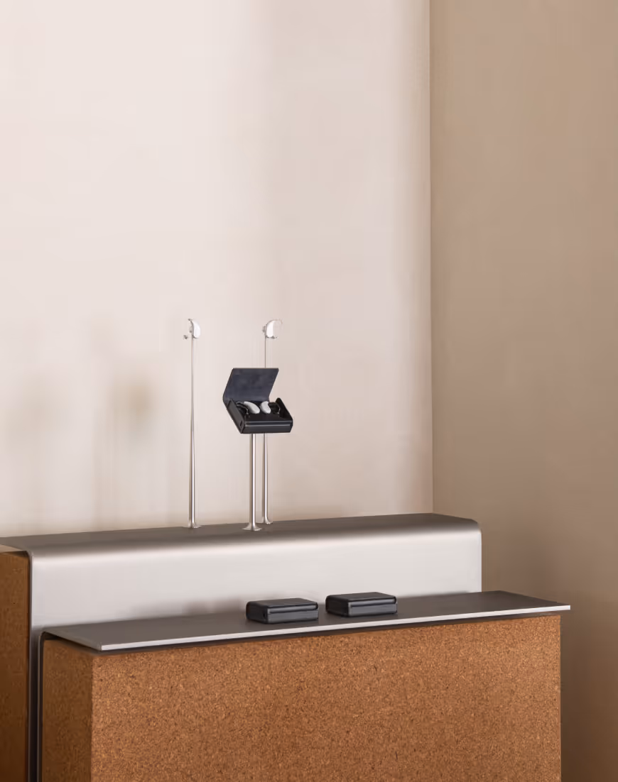 Two pairs of white hearing aids displayed on metal stands and two black carrying cases on a cork and gray surface against a beige wall.