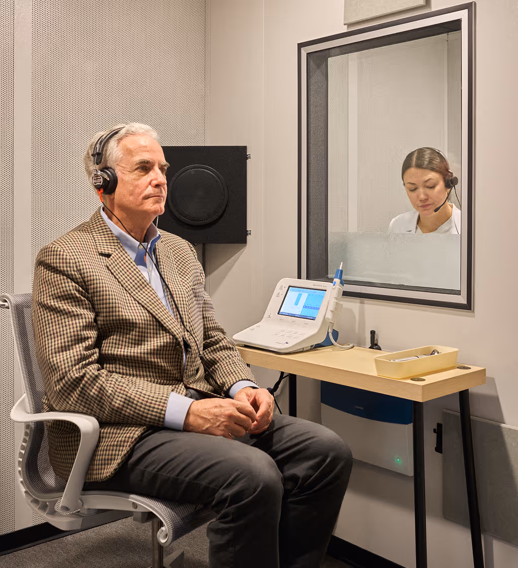 Man wearing headphones sitting in a soundproof booth during a hearing test, with a woman audiologist observing through a glass window.