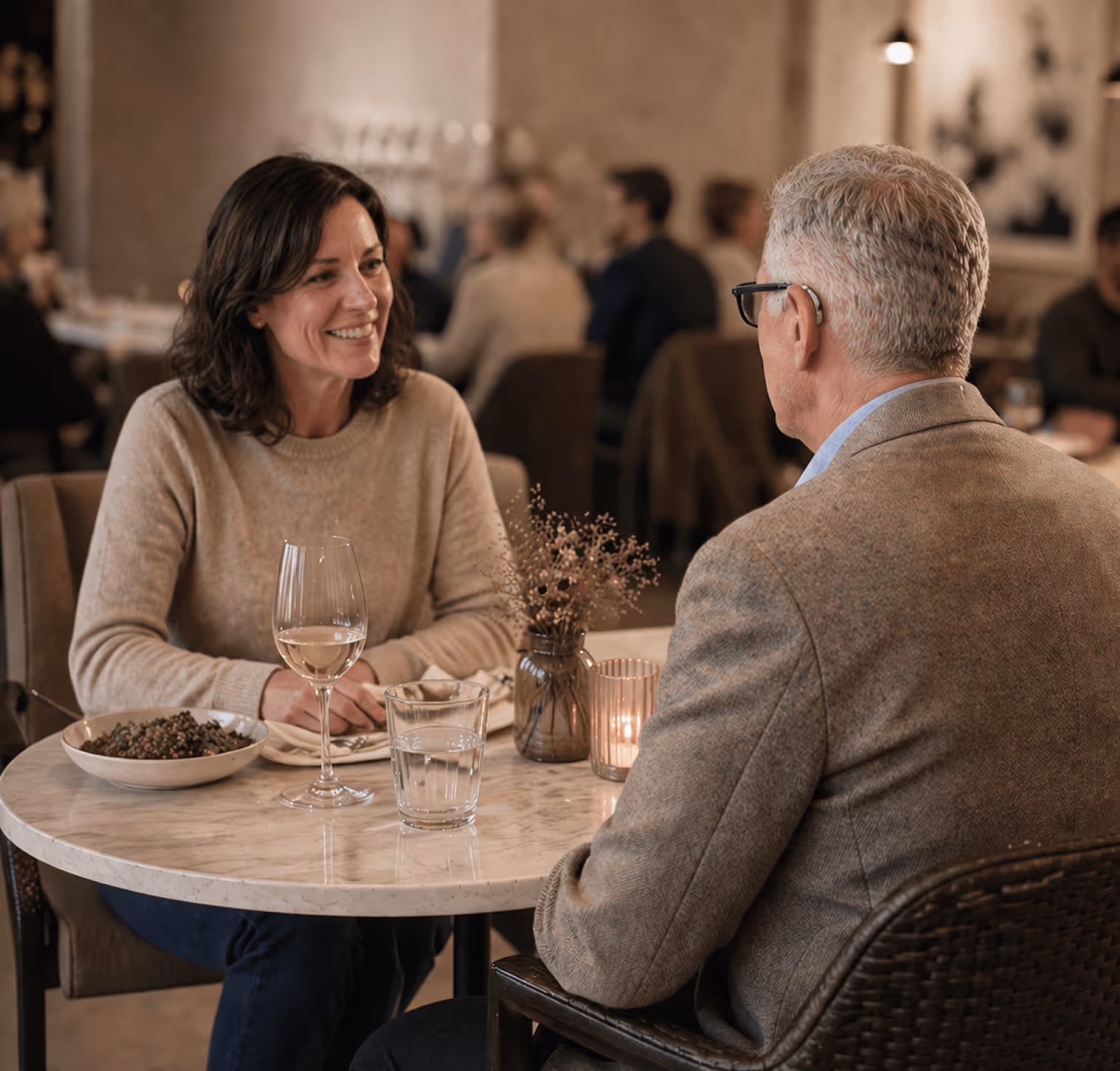 Smiling woman and older man sitting at a marble table in a restaurant, with wine glass, water glass, and a bowl of food on the table.