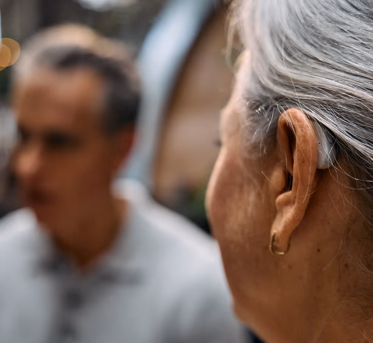 Close-up of an elderly woman with gray hair wearing a hearing aid, facing a blurred man in the background.