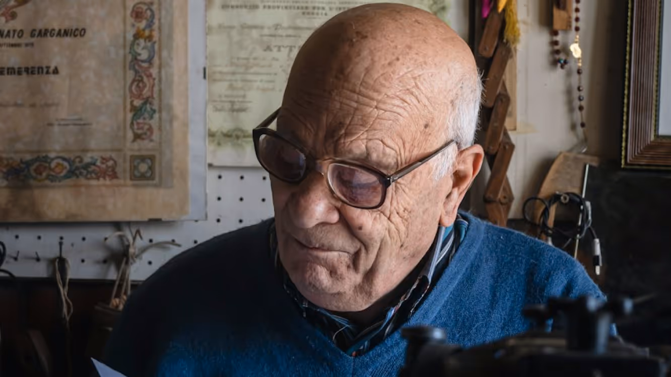 Elderly man wearing glasses and a blue sweater looking down, with certificates and various items on the wall behind him.