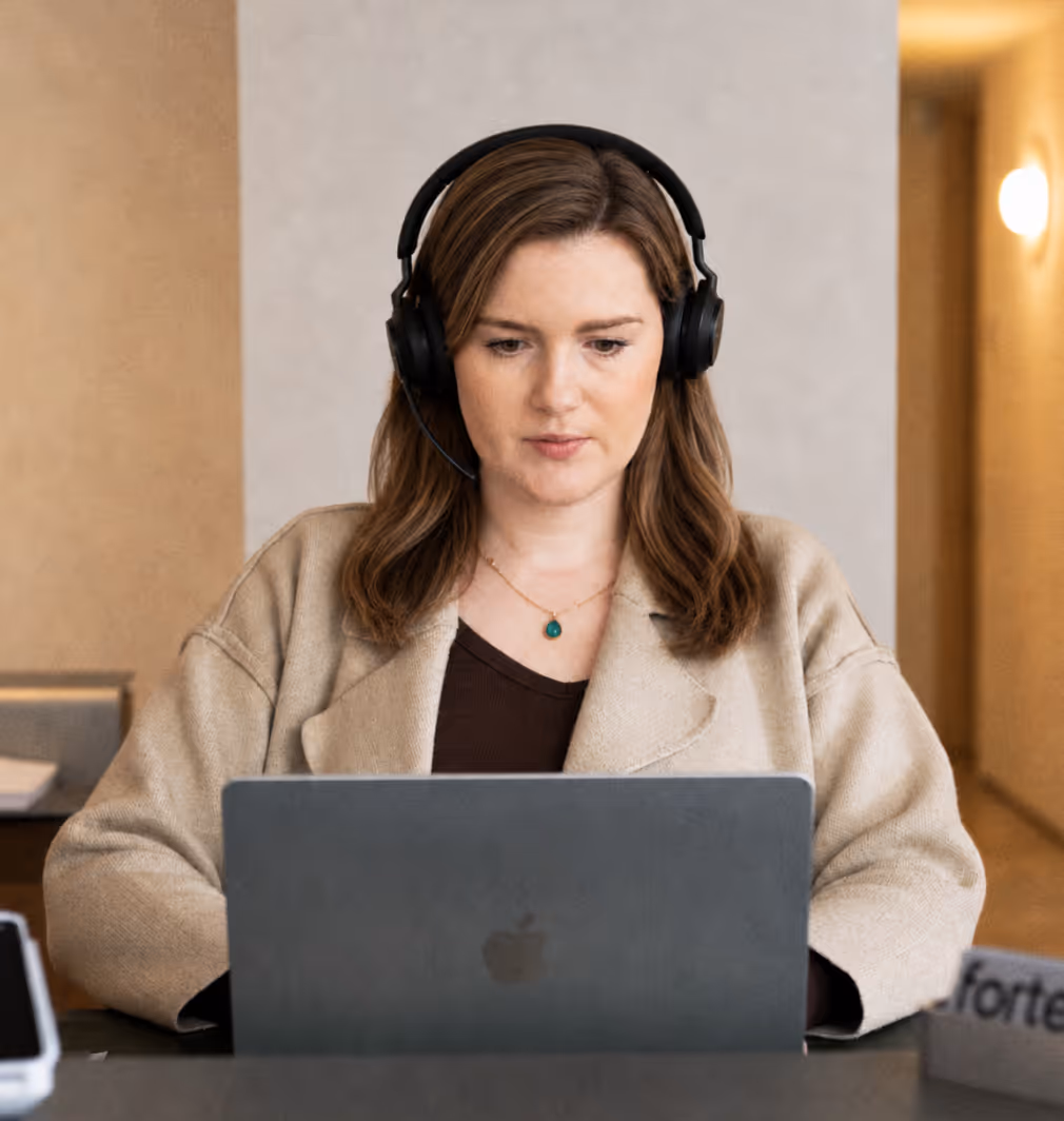Woman wearing headset and beige jacket focused on working on a laptop.