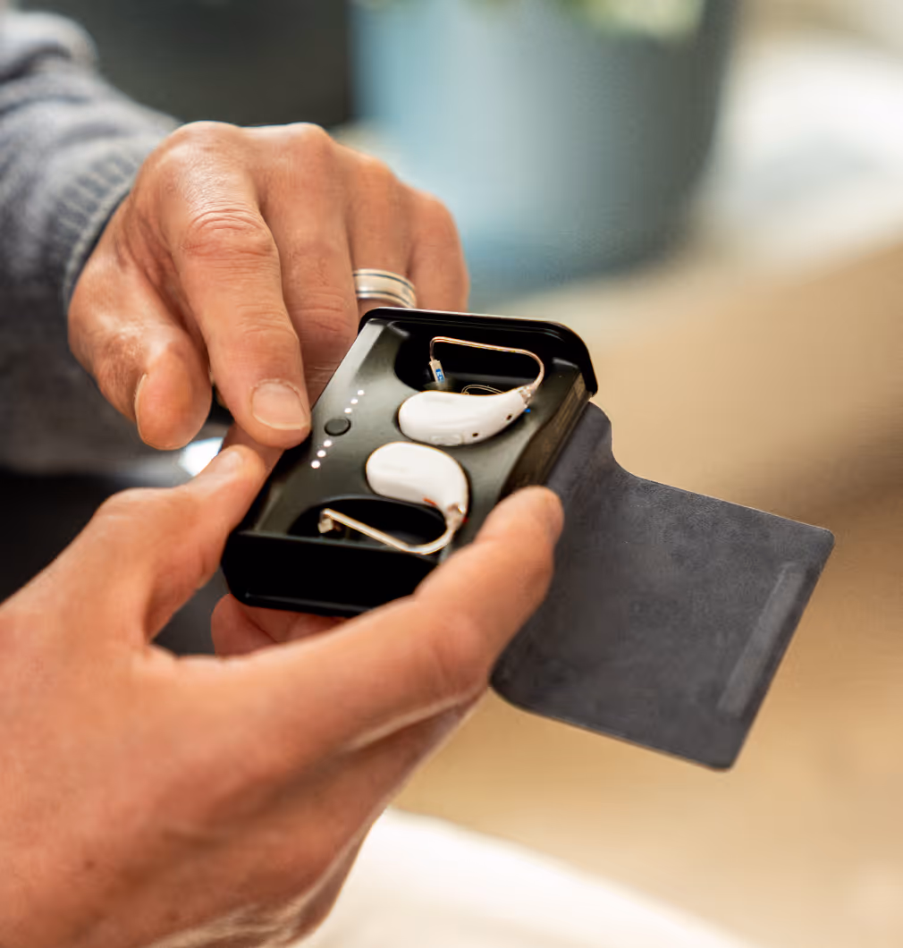 Two hands holding a black charging case with white hearing aids inside and LED charge indicators.