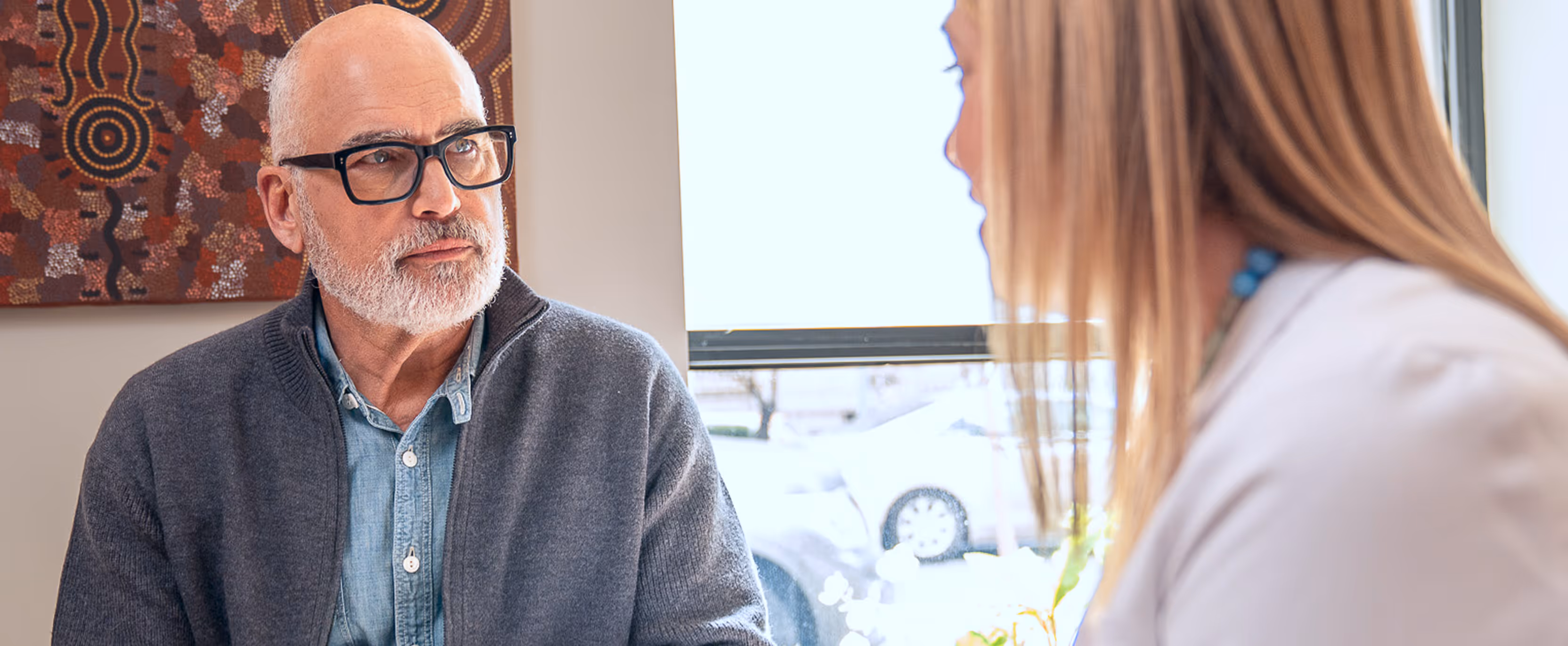 Older man with glasses and gray beard listening attentively to a woman in a white coat.