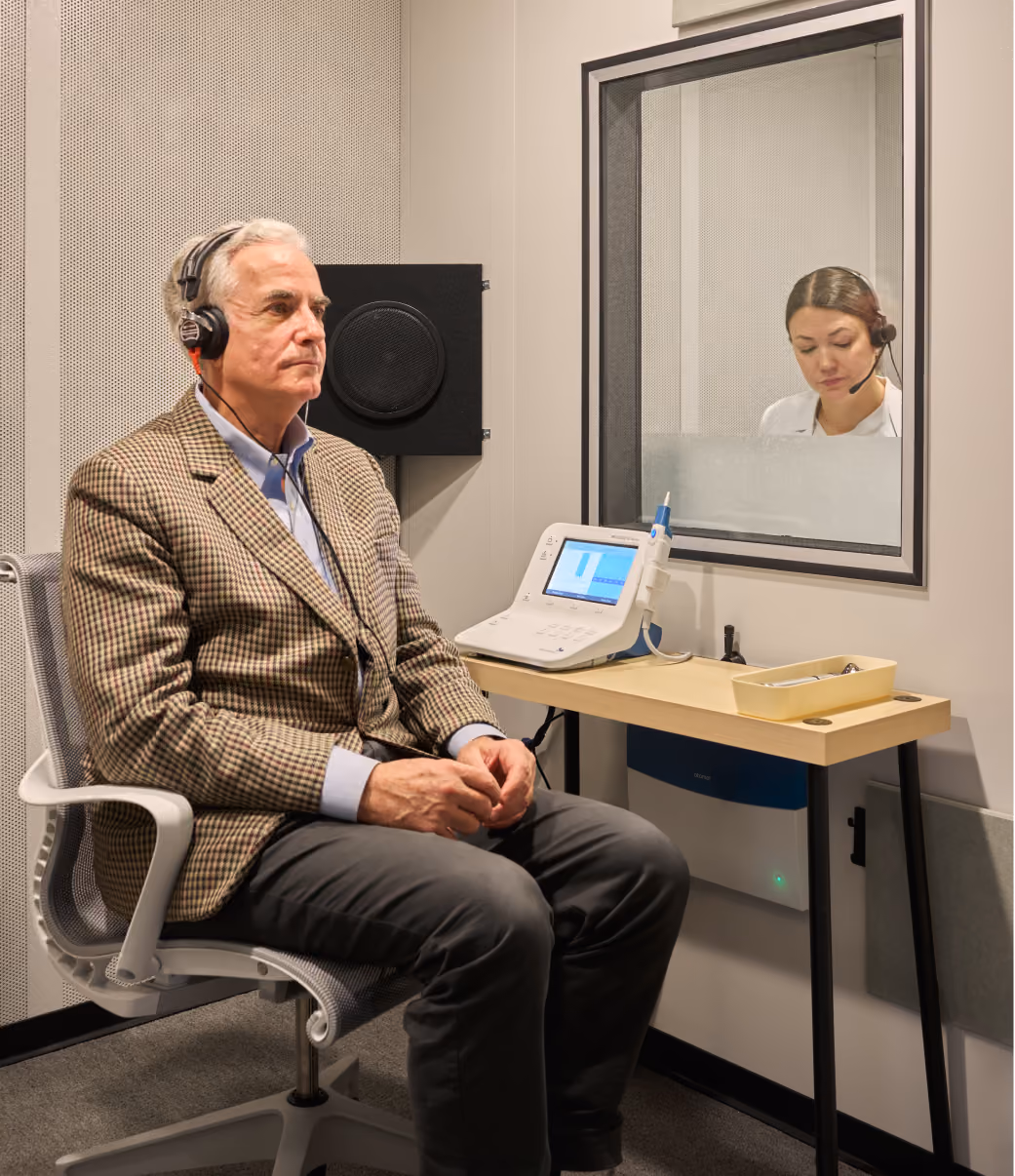 Patient in a sound booth taking a hearing test while an audiologist watches through the window.