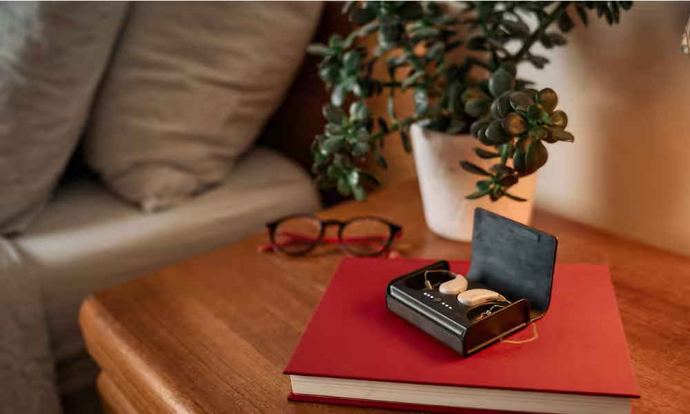 Fortell hearing aids in their open black charging case on a red book, beside glasses and a houseplant.