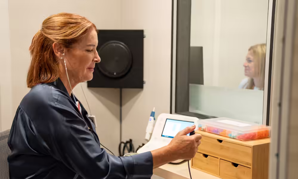 Woman wearing insert earphones presses a response button in a sound booth while an audiologist watches through the window.