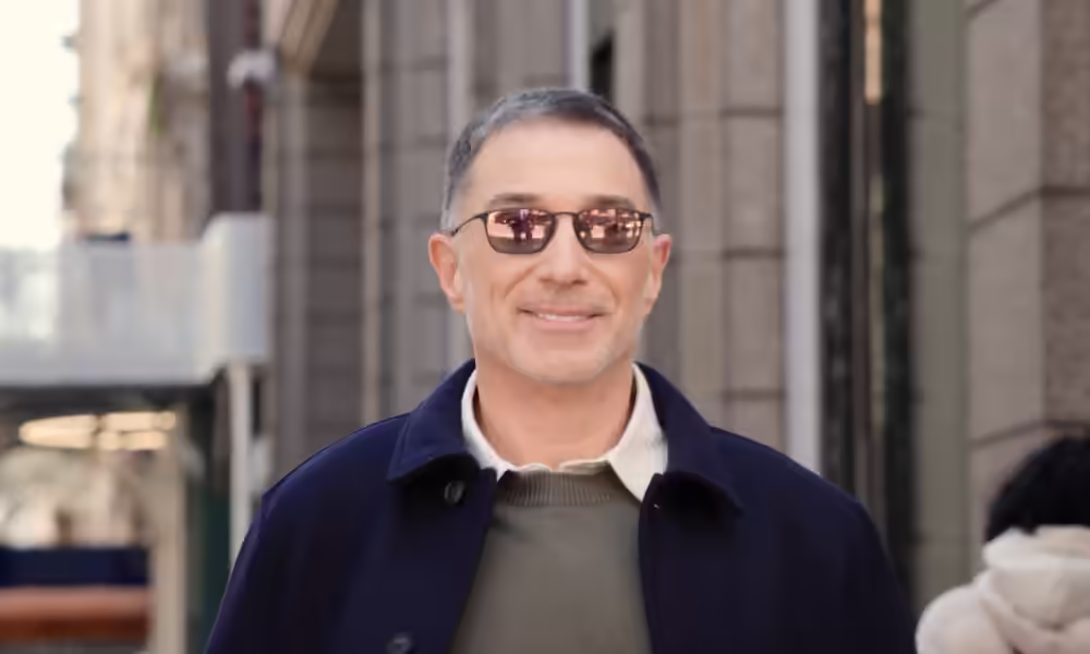 Smiling man in sunglasses and a navy coat on a city street.