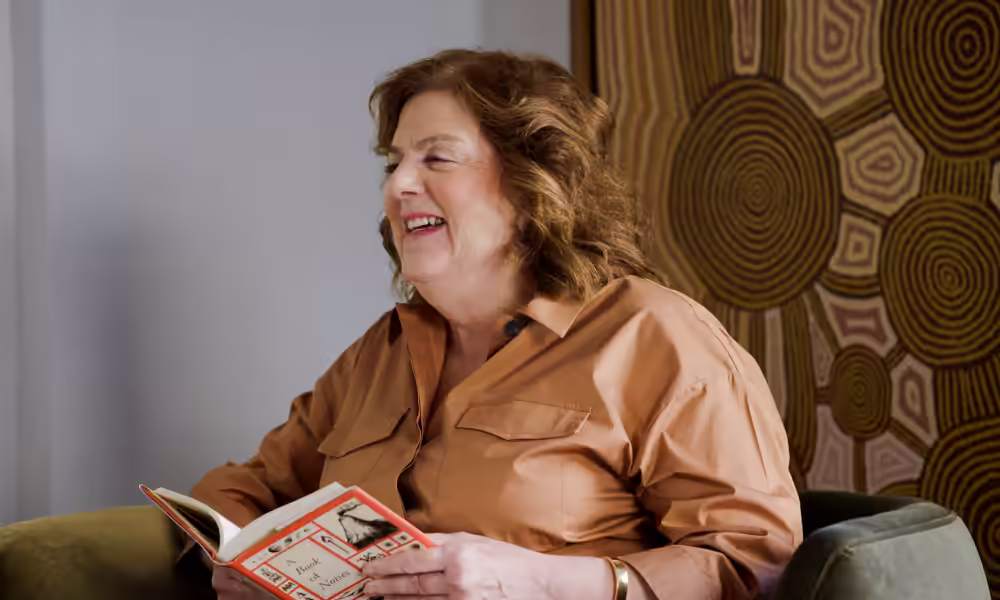 Older woman laughing in an armchair while holding an open book.