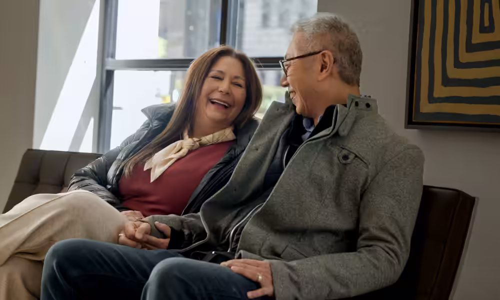 Older couple laughing together on a leather sofa by a sunny window.