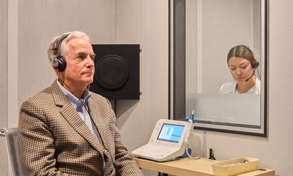 Older man wearing headphones in a sound booth while an audiologist runs the test from behind a window.