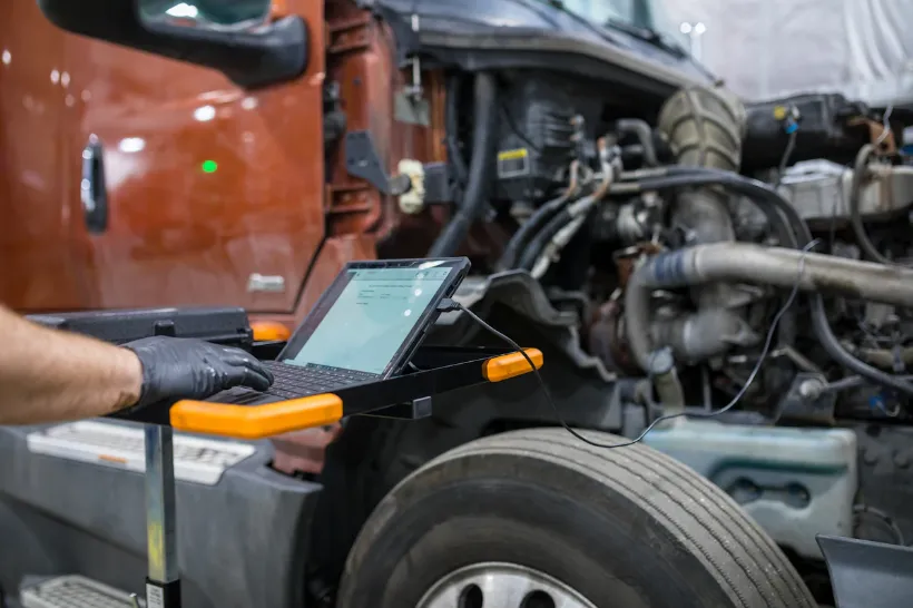 Heavy-duty diagnostic service, technician using laptop to scan truck engine with hood open in shop.