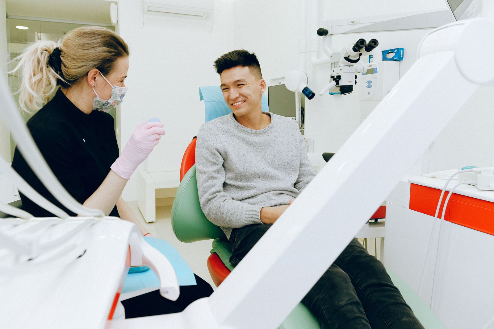 A dental professional in black scrubs talks with a smiling patient in a modern operatory, a microscope and delivery unit visible beside the chair.
