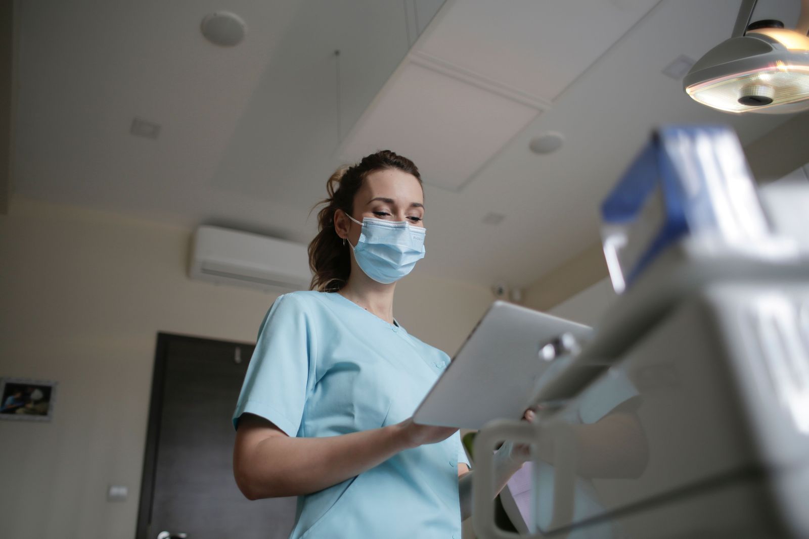 A dental professional in light blue scrubs and a mask looks at a tablet in a bright operatory, with the exam light visible above.