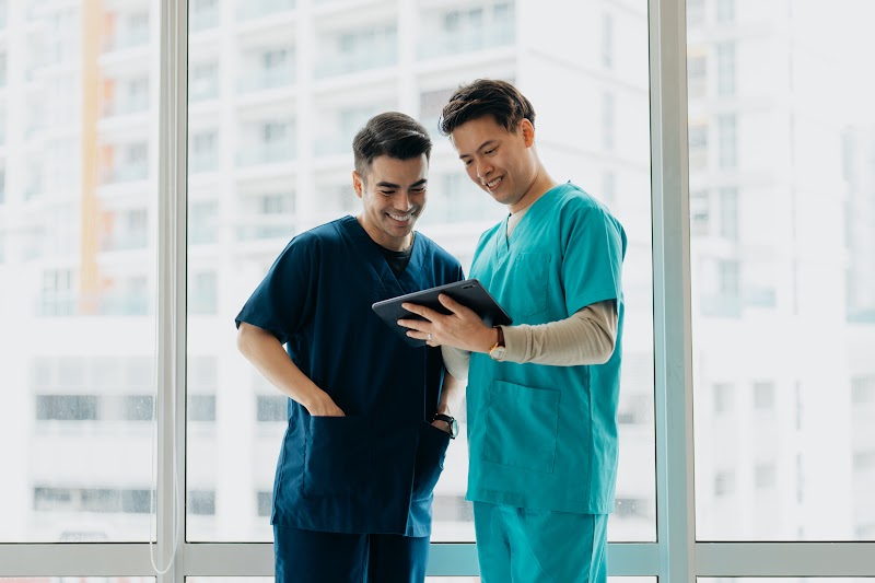 Two dental professionals in navy and teal scrubs stand by a bright window and look at a tablet together.