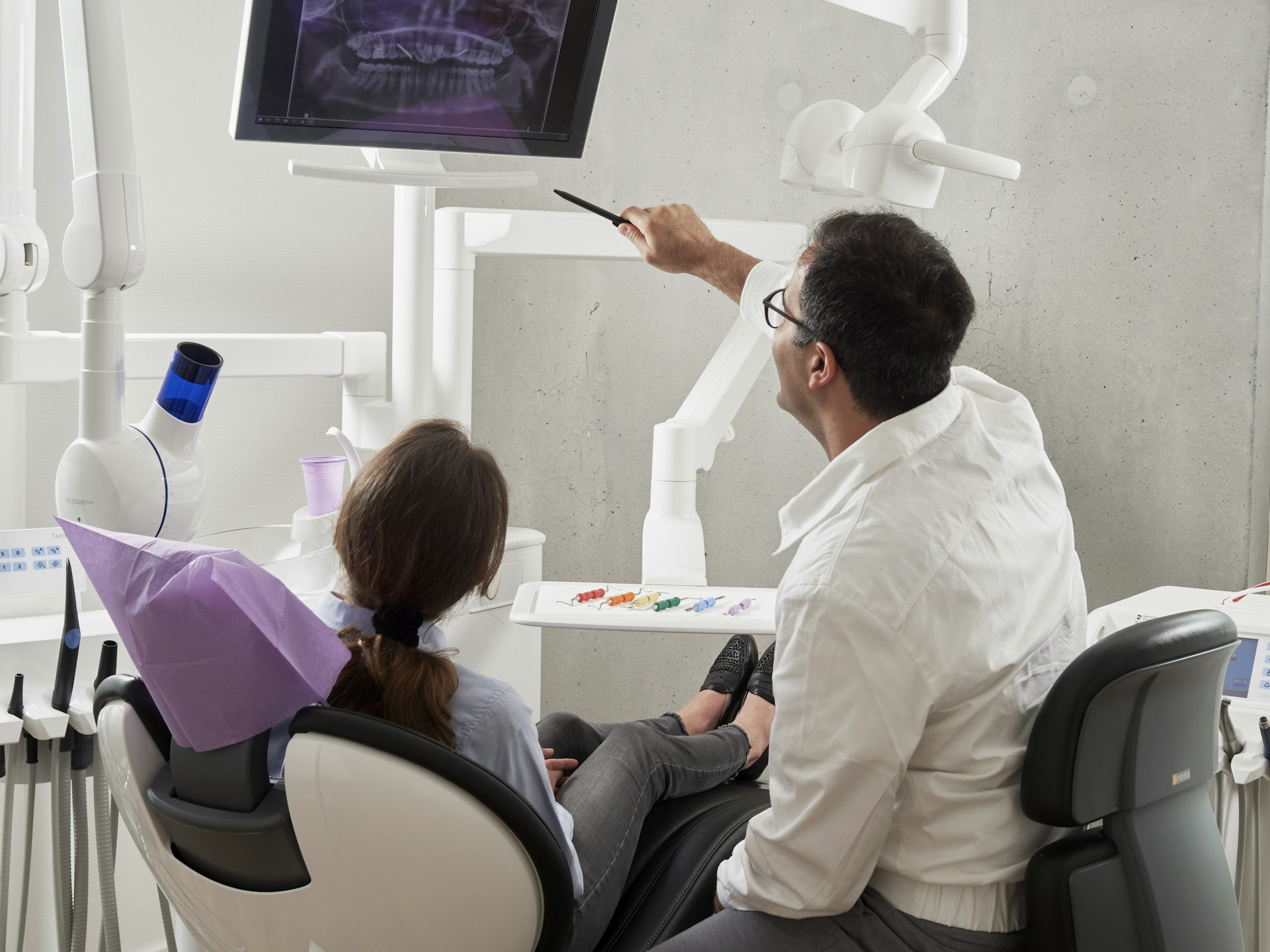 A dental professional in a white clinic jacket points at a panoramic X-ray on a monitor while a patient in a bib looks on from the chair.