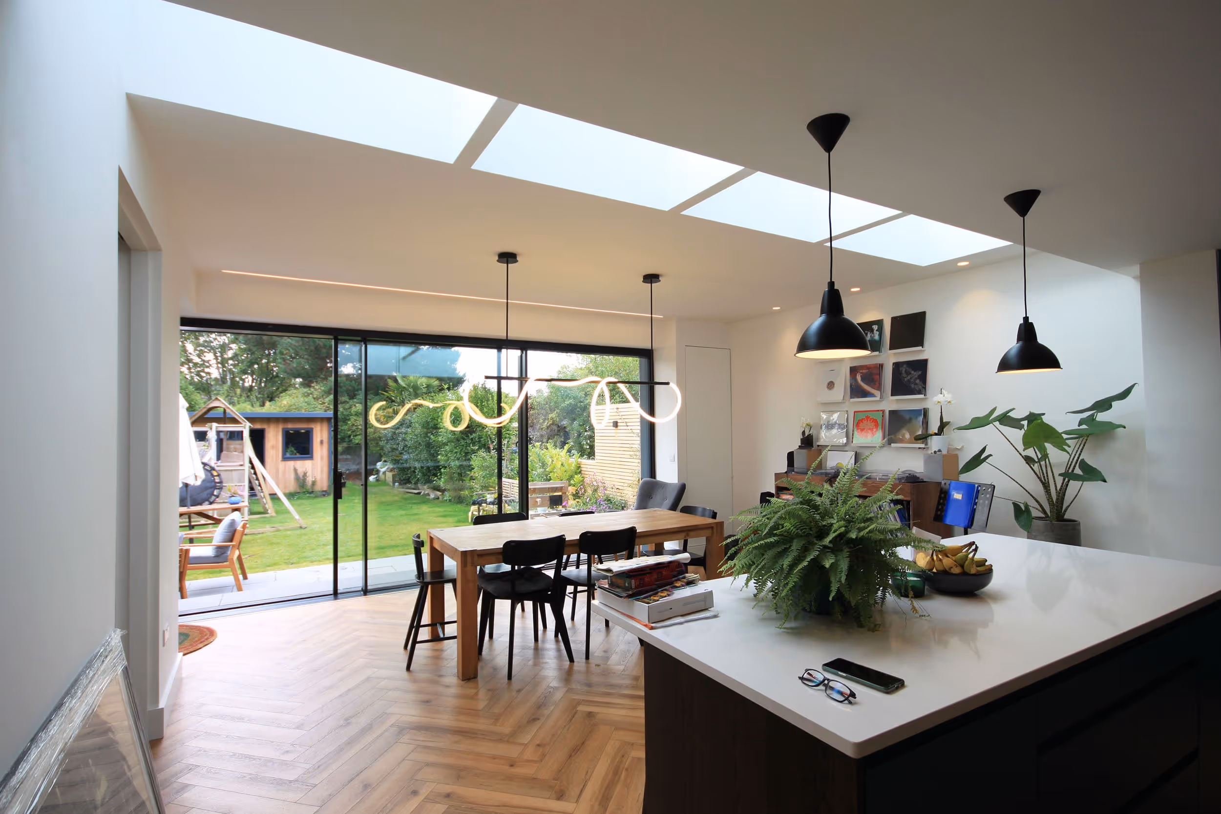Interior shot of a kitchen and dining room with a kitchen island in the middle and dining table at the back, big windows looking onto garden and skylights