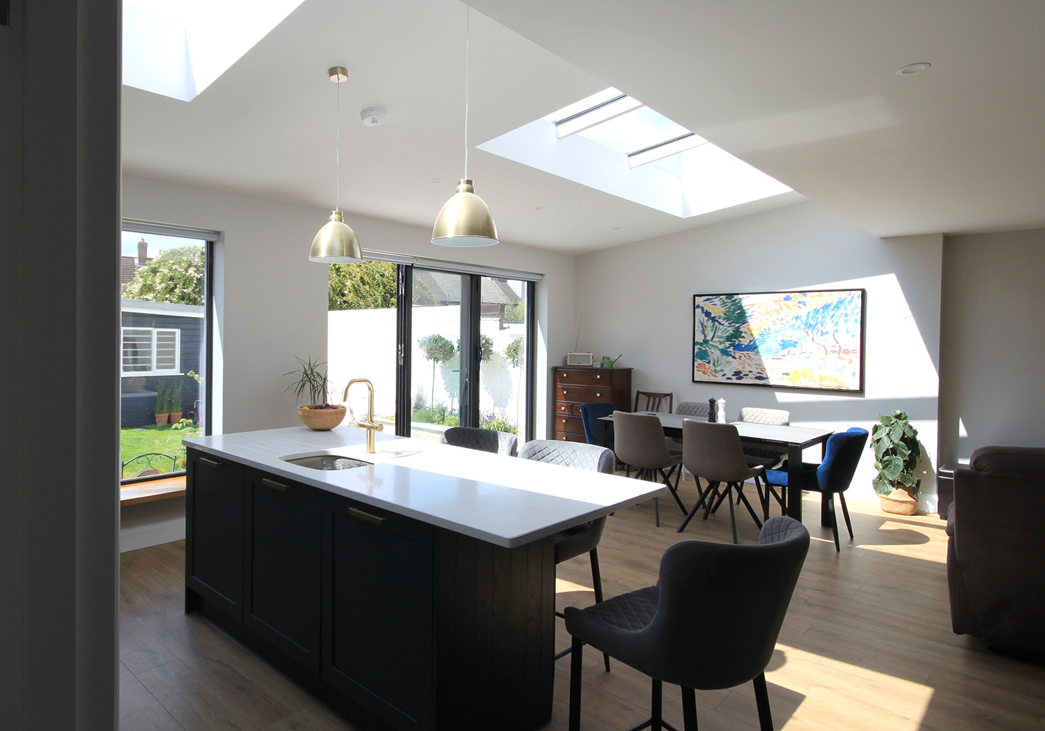 Interior shot of a kitchen and dining room with a kitchen island in the middle and dining table at the back, big windows looking onto garden and skylights