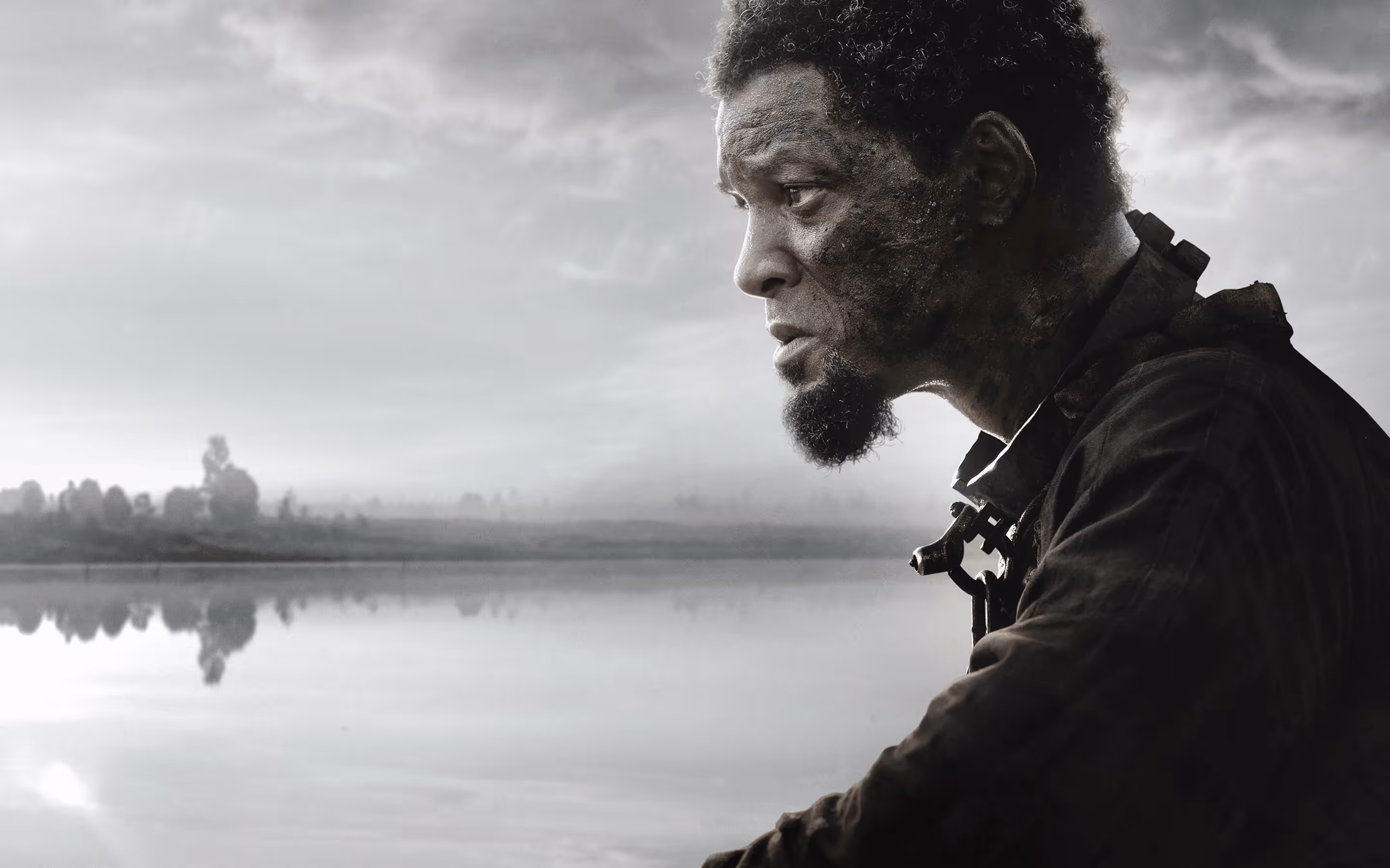 Side profile of a man with a textured beard and worn clothing, looking thoughtfully across a calm lake with a cloudy sky.