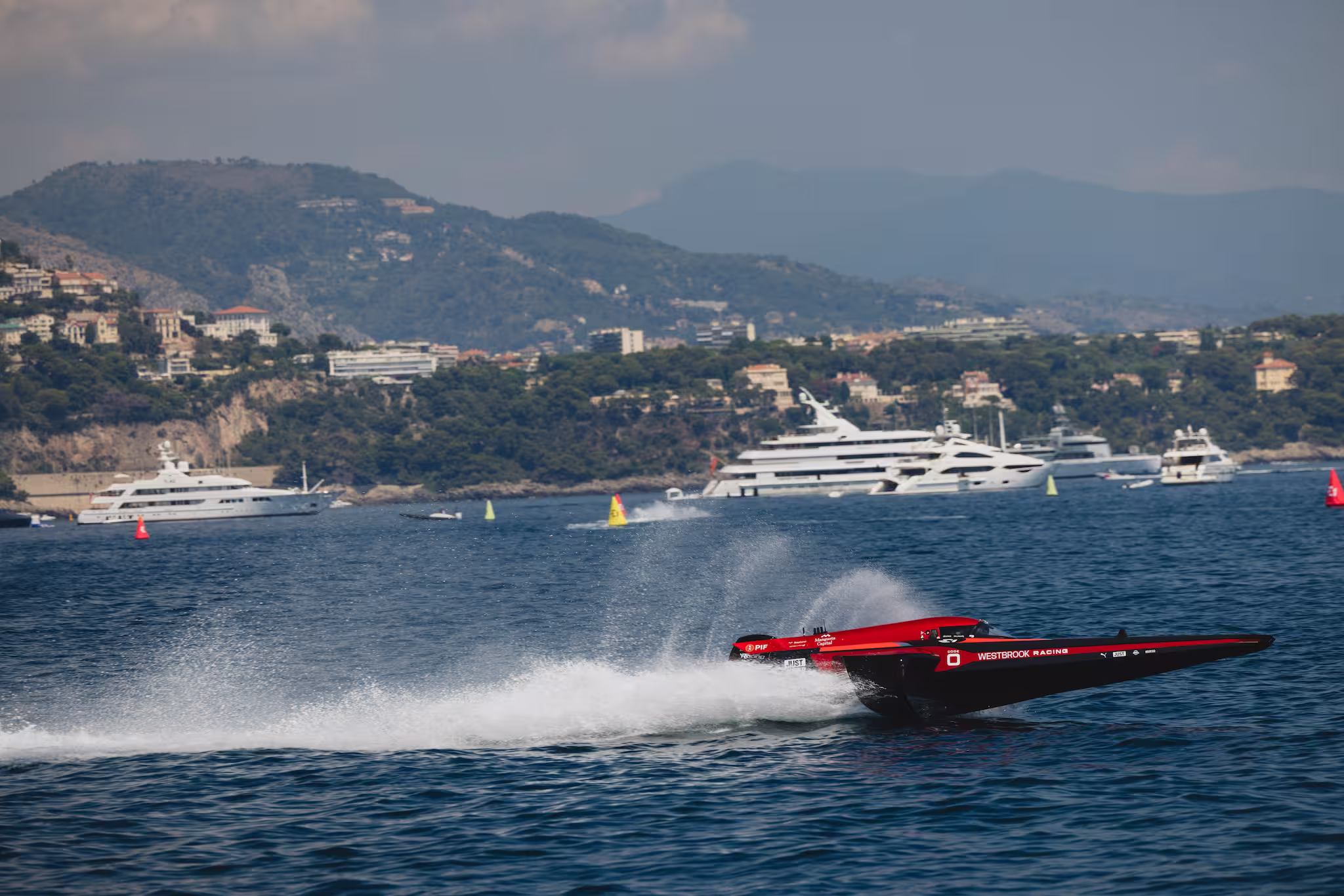 A red and black Westbrook Racing powerboat speeding across a blue sea with yachts and hills in the background.