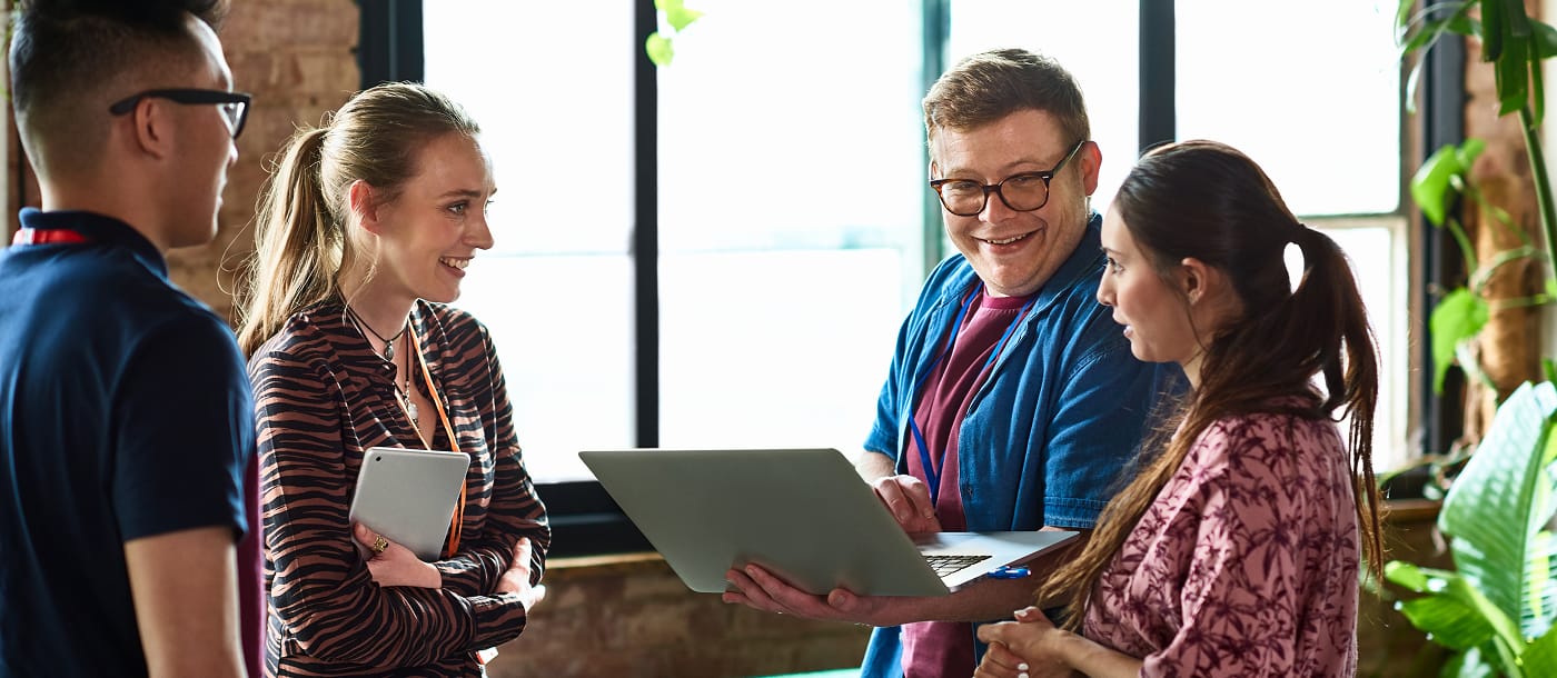 Four young professionals having a discussion in an office, one holding a laptop and another holding a tablet.