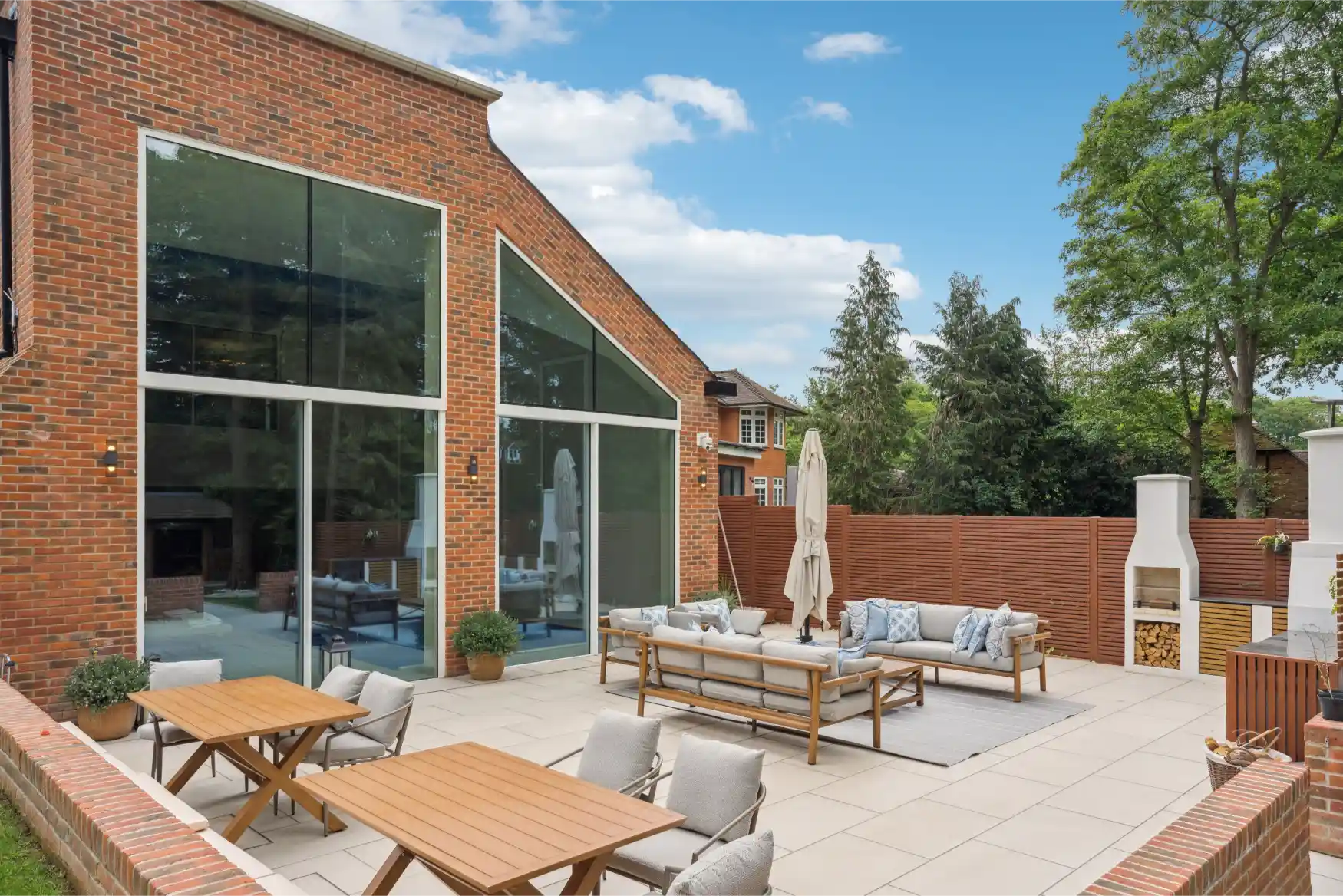 Spacious patio with brick house, large glass windows, outdoor seating, wooden tables, and garden view.