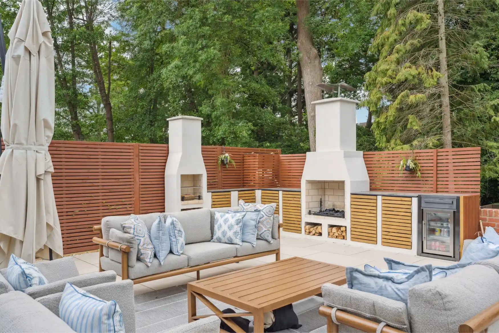 Outdoor patio with modern sofas, wooden table, built-in fireplaces, mini fridge, and privacy fence.