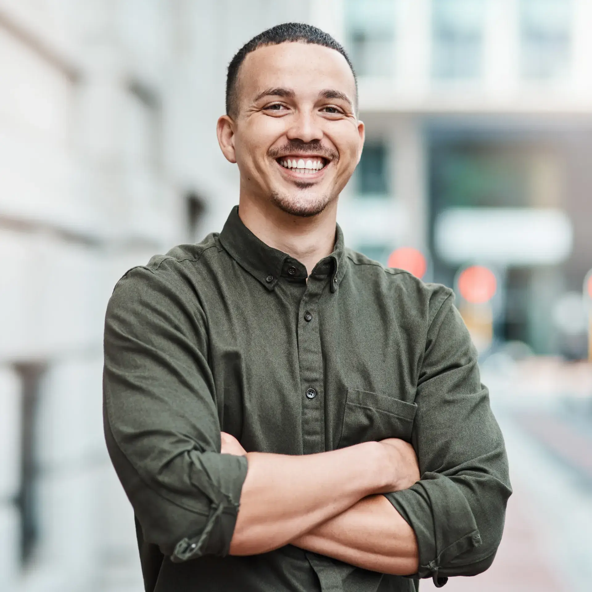 A man stands outside, smiling with arms crossed, wearing a green button-up shirt.