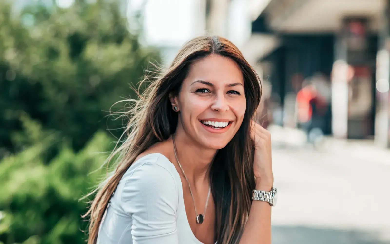 A woman with long hair smiles while sitting outdoors, wearing a white shirt and watch.