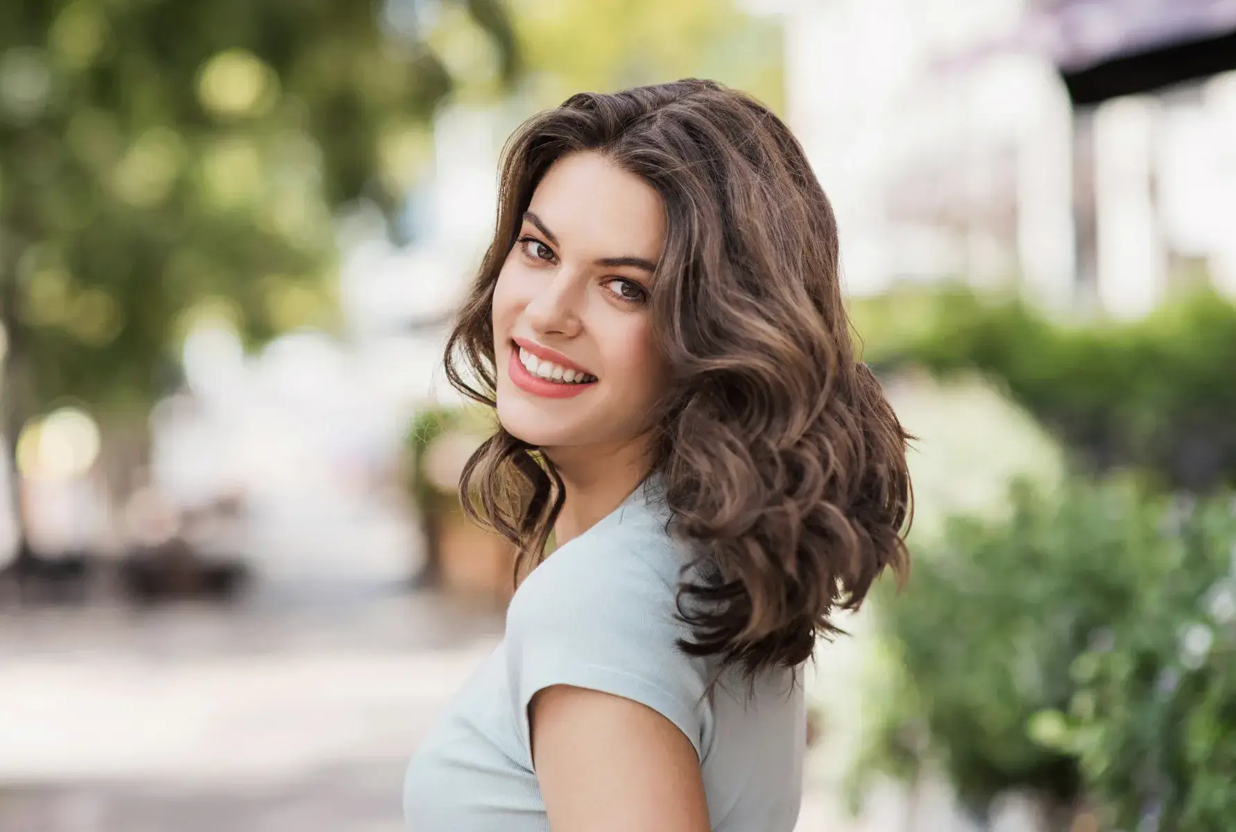 Smiling woman with wavy hair looks over her shoulder outdoors on a sunny day.