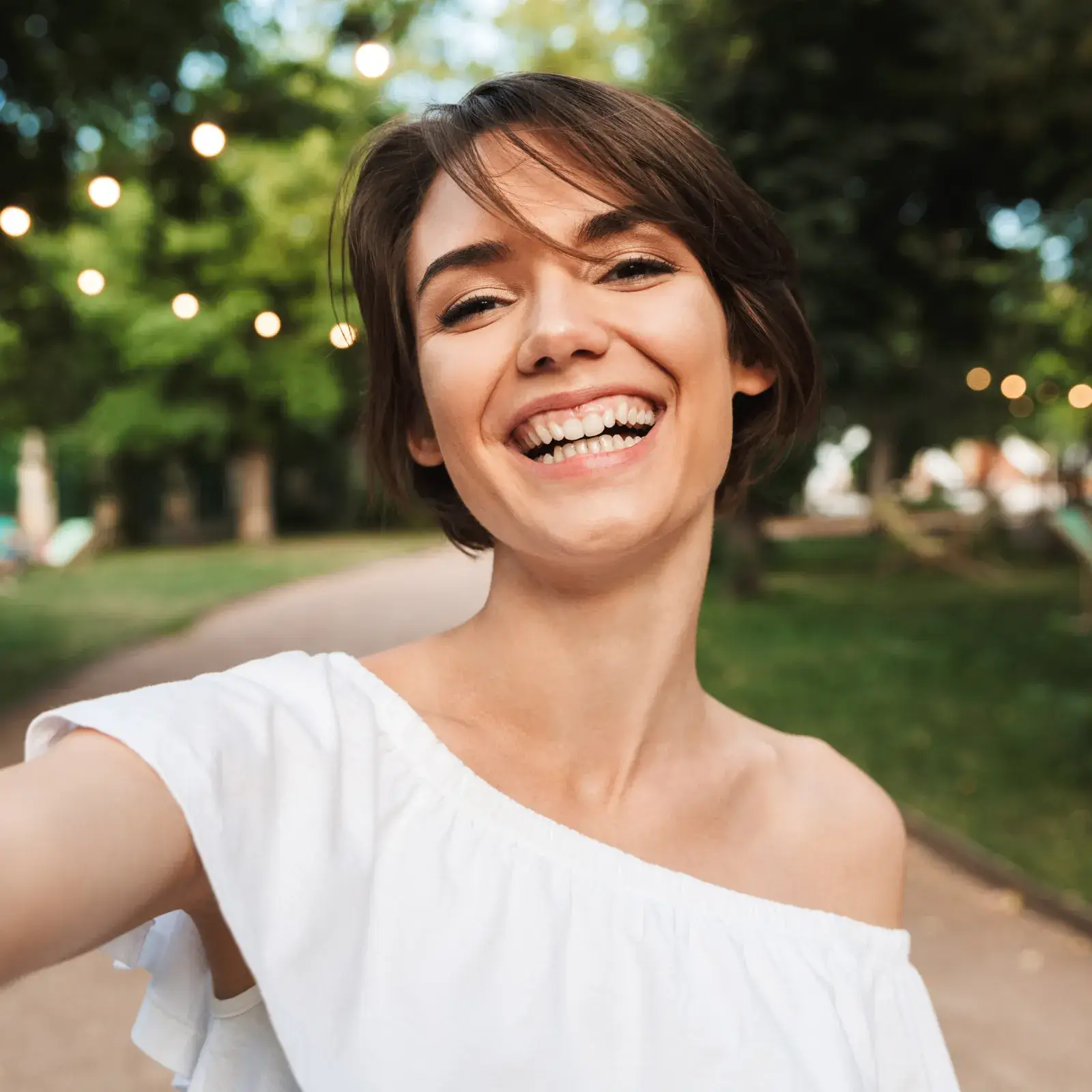 Woman smiling widely while taking a selfie outdoors in a park.