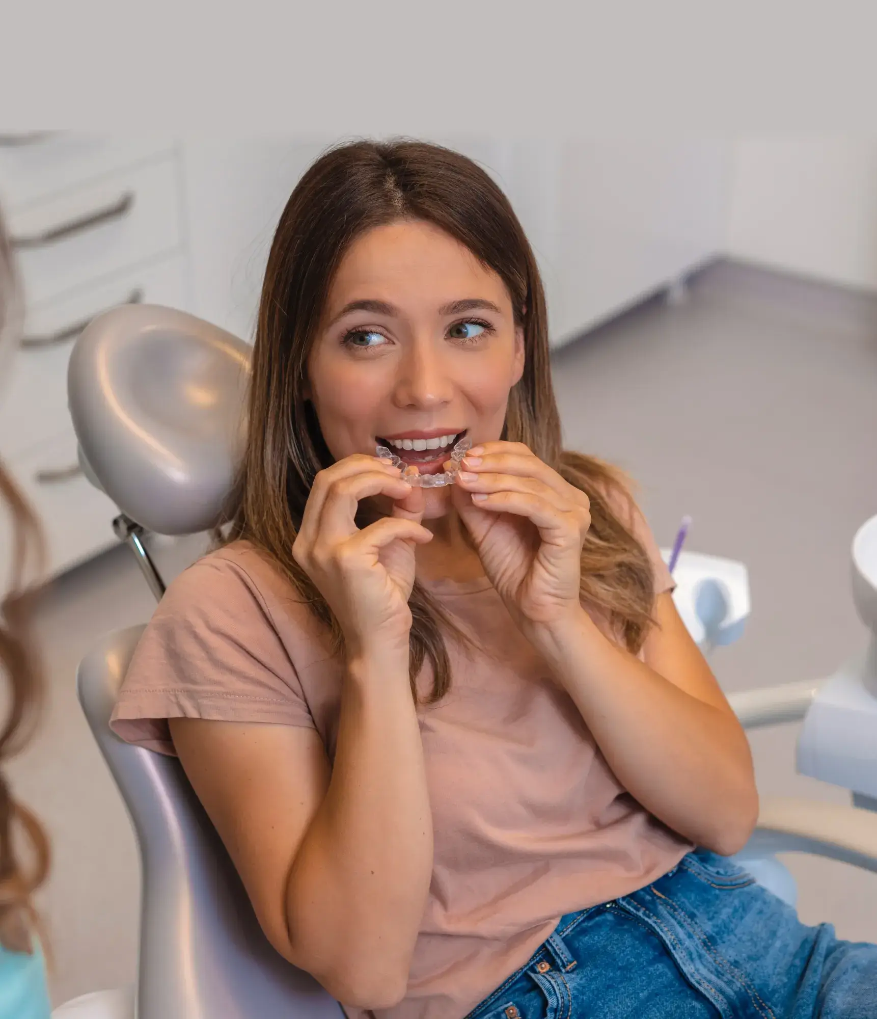 A woman sitting in a dental chair inserts a clear retainer into her mouth.