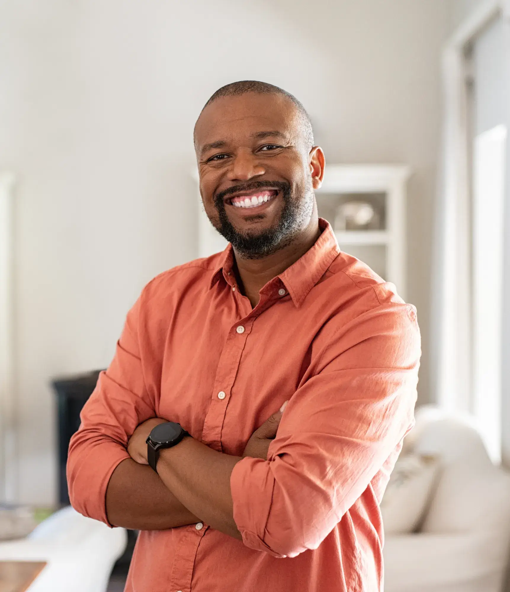 A man in a red shirt smiles with crossed arms in a bright room.