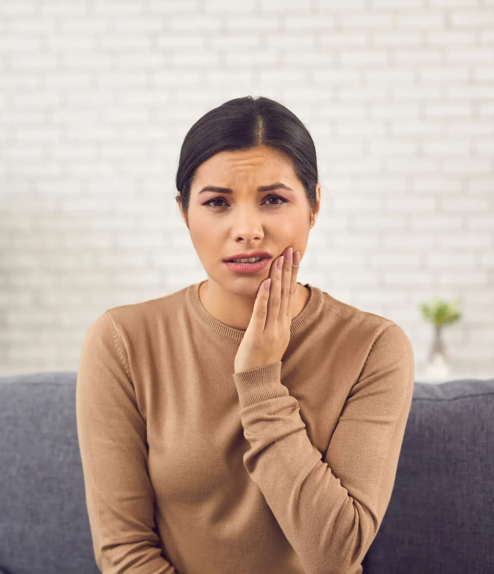 A woman sitting on a couch, holding her cheek in pain, possibly due to a toothache.
