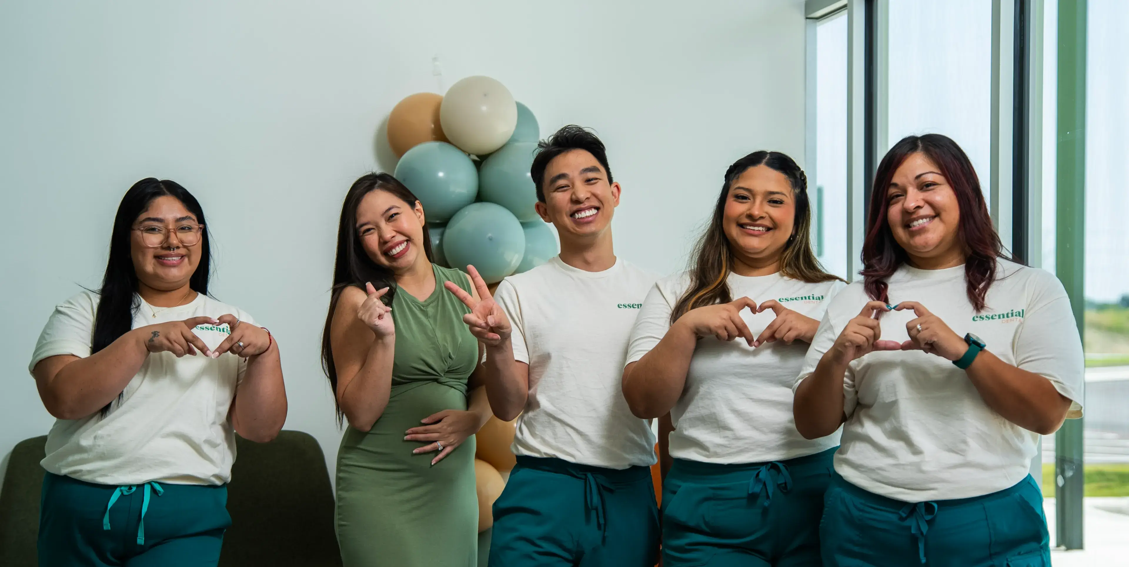 Five people stand smiling, making heart gestures with their hands in a bright room.