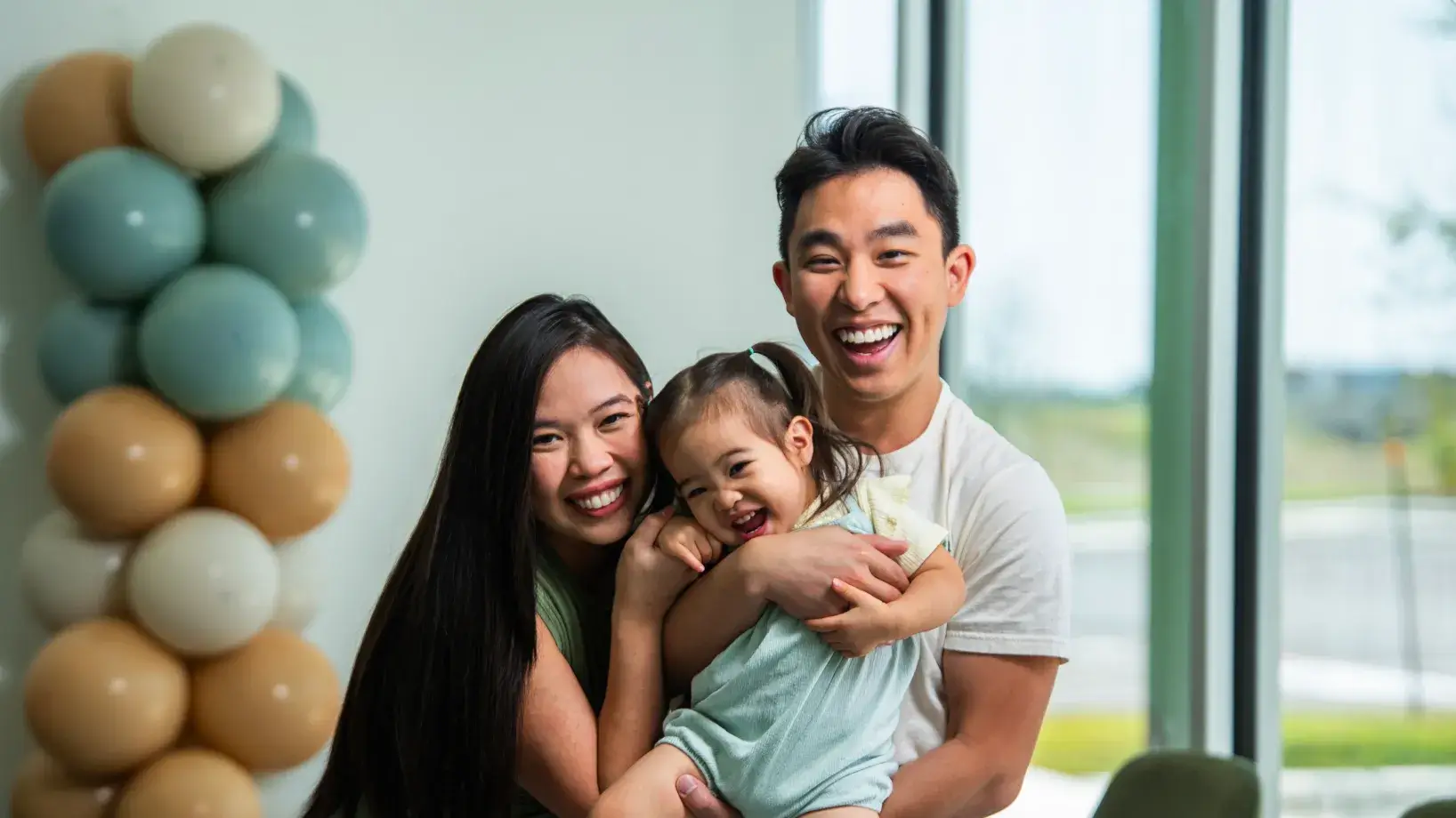 A smiling couple holds their young child in their arms indoors.