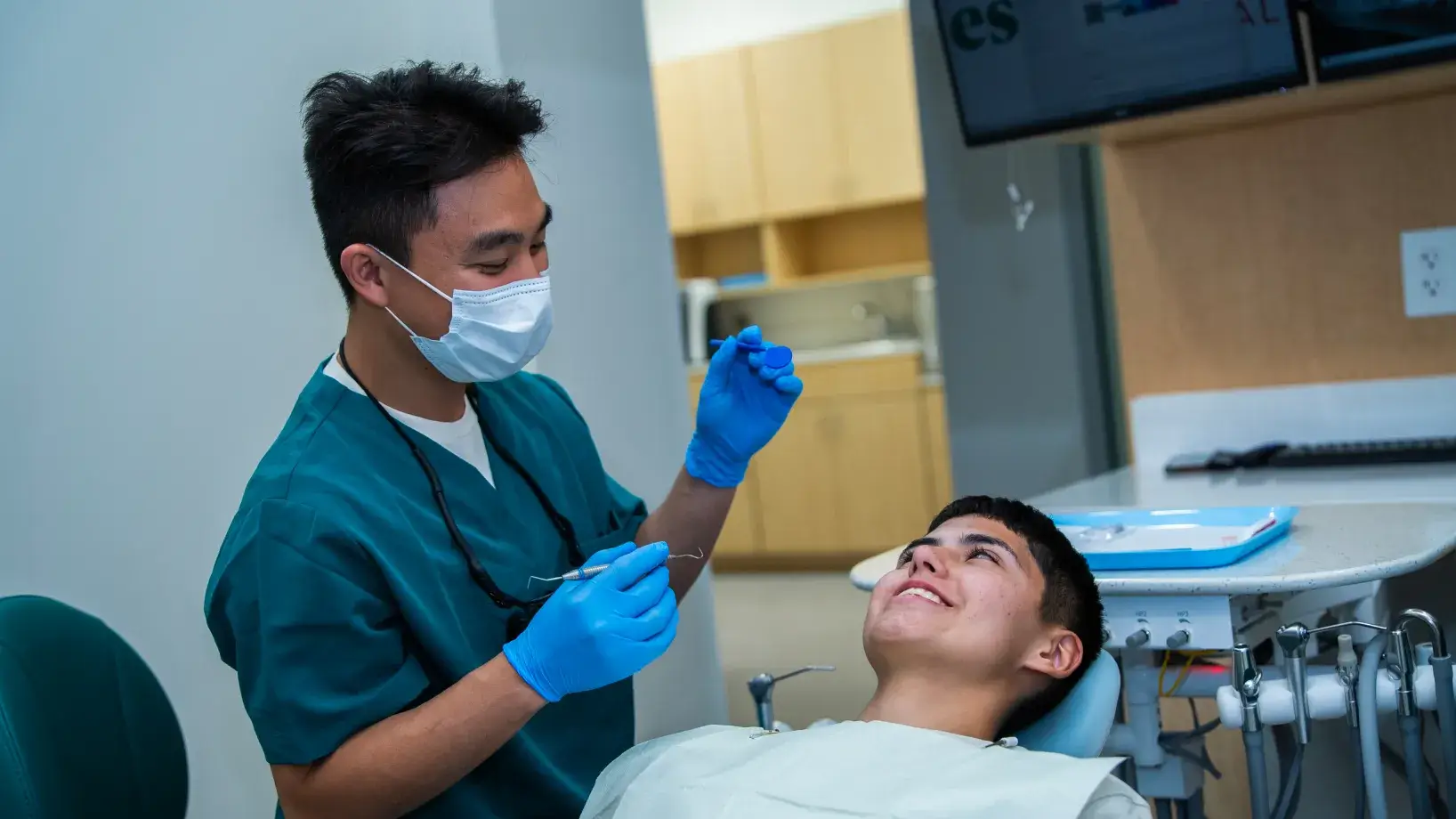 A dentist in green scrubs examines a smiling patient in a dental chair.