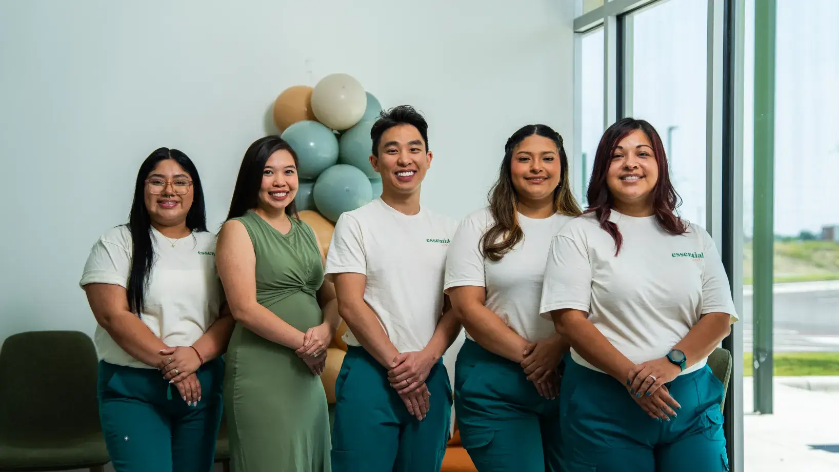A group of five people smiling, standing indoors near a wall with balloons.