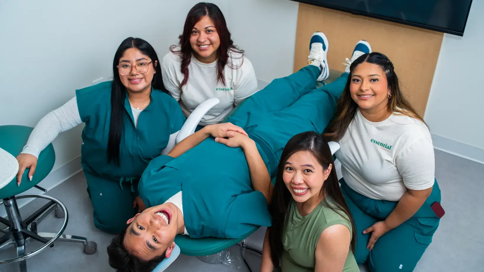 A person lies on a dental chair surrounded by four smiling people in an office setting.