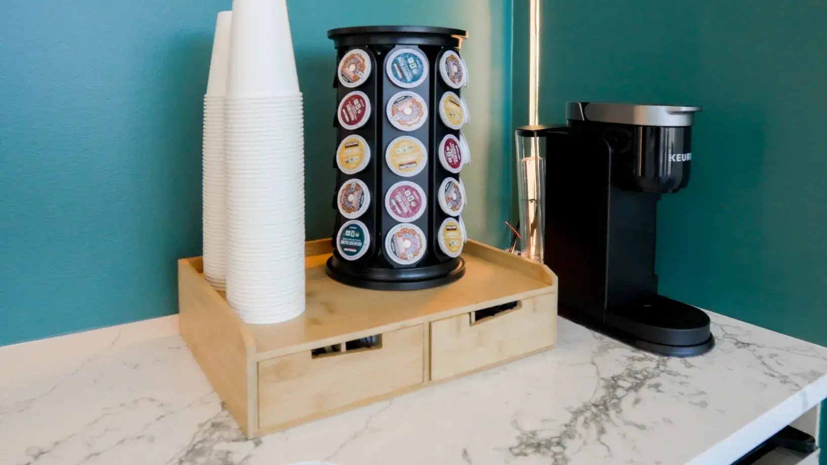 A coffee station with a Keurig machine, a carousel of coffee pods, and stacked paper cups.