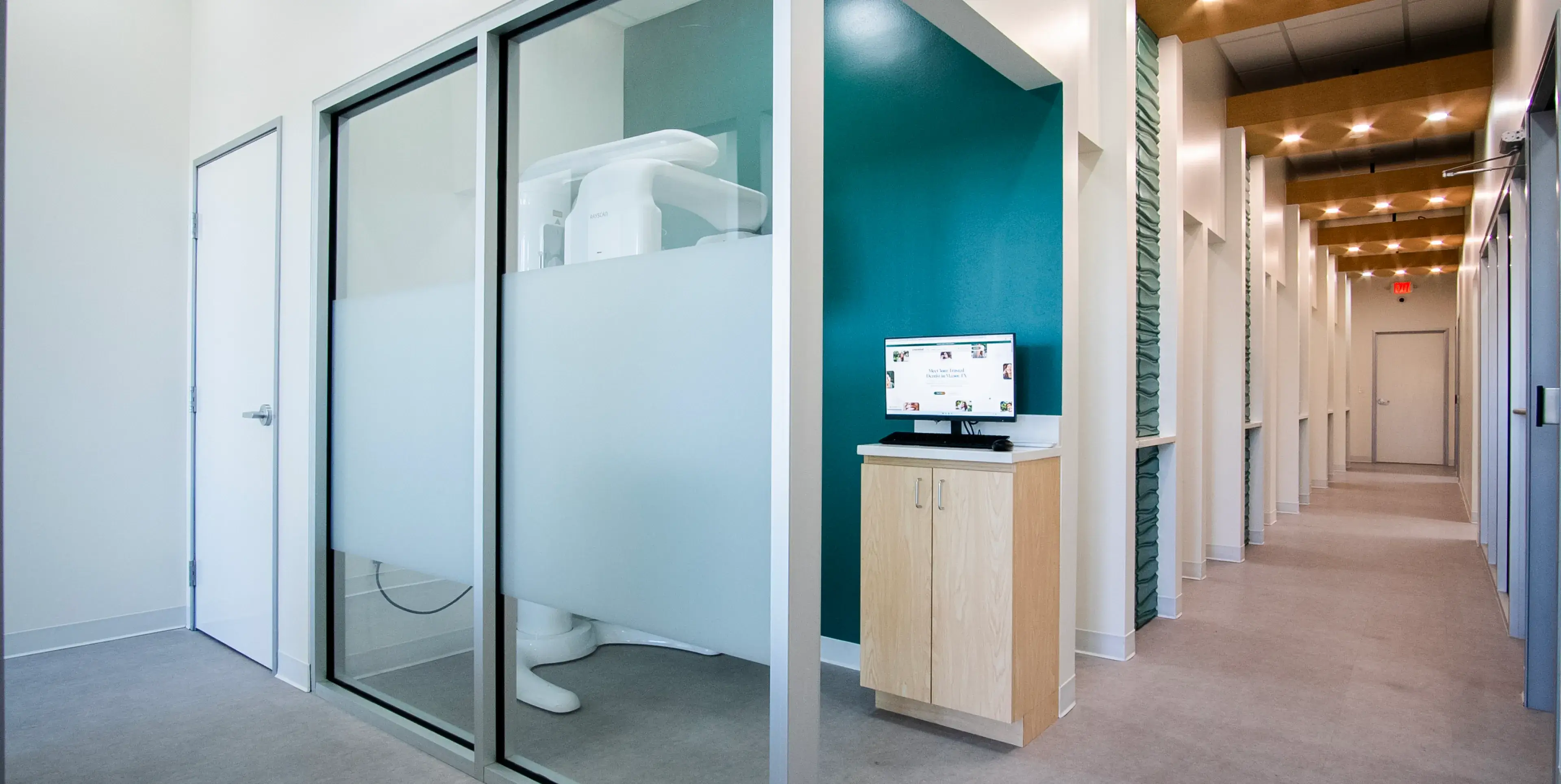 Dental office hallway with a digital screen on a wooden cabinet beside a frosted glass room.