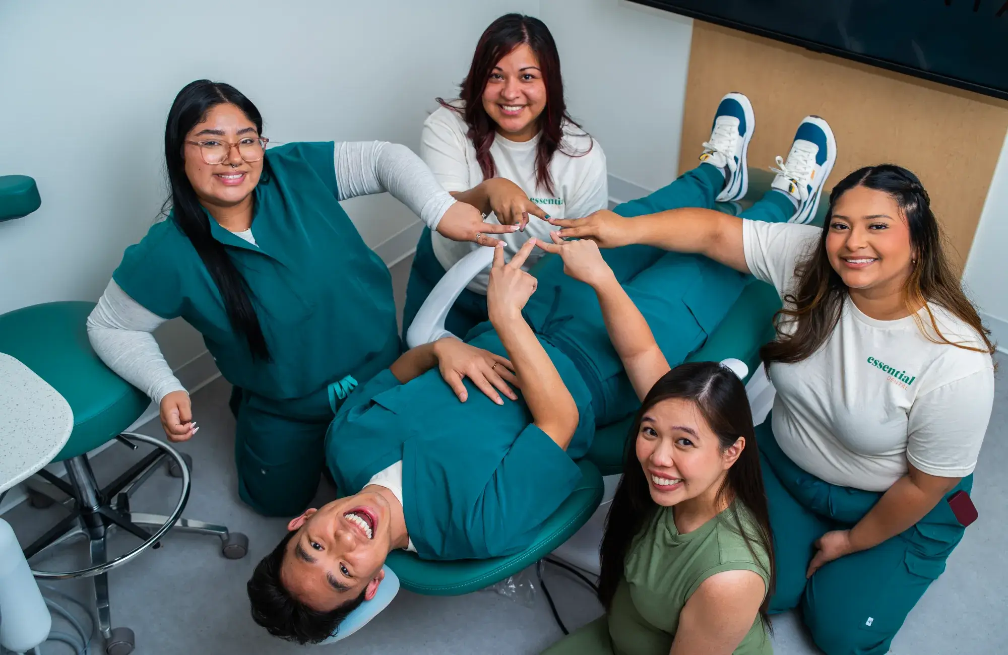 Five dental professionals smile while playfully posing around a patient reclining in a dental chair.