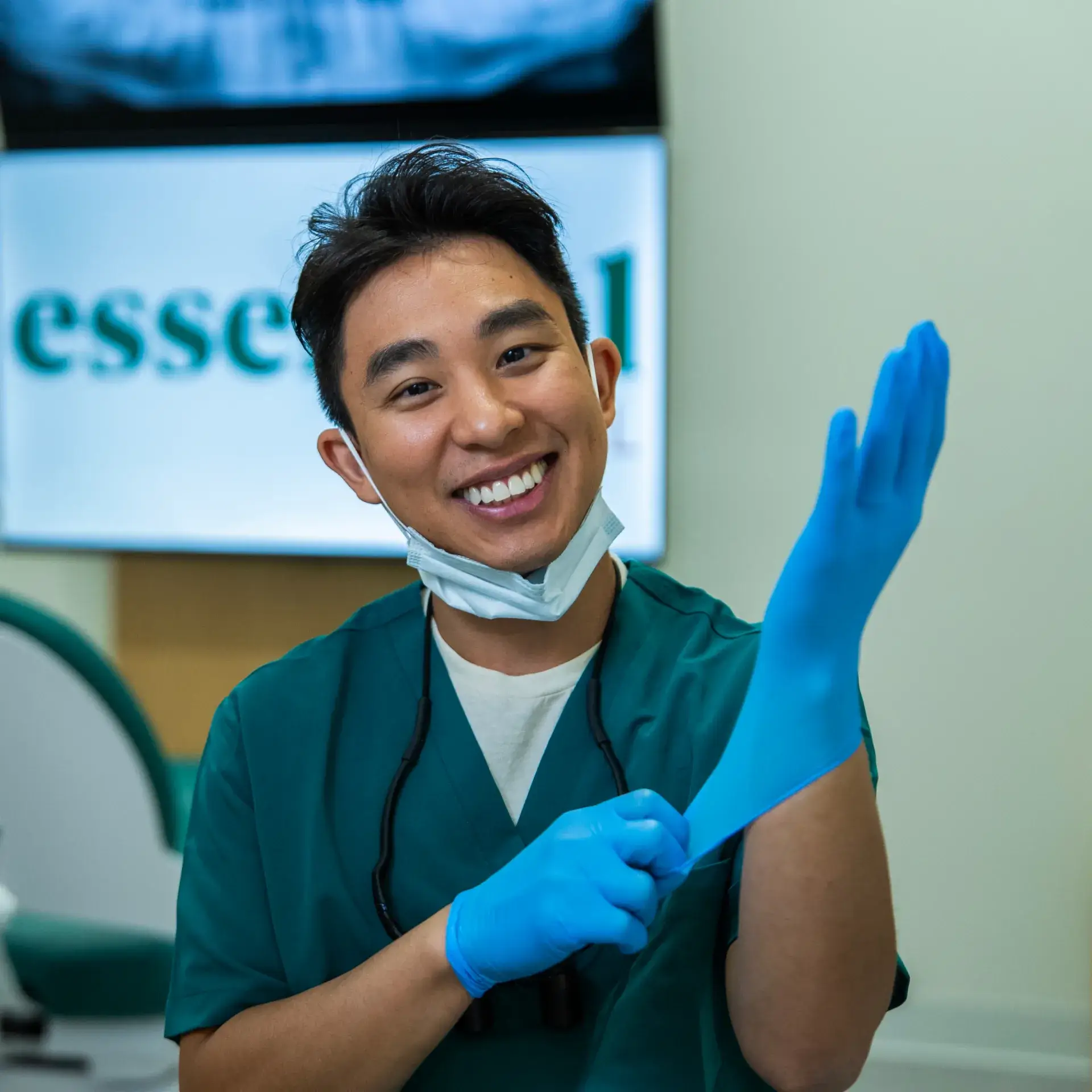 A person in a dental clinic putting on blue gloves, smiling, and wearing a mask around their neck.