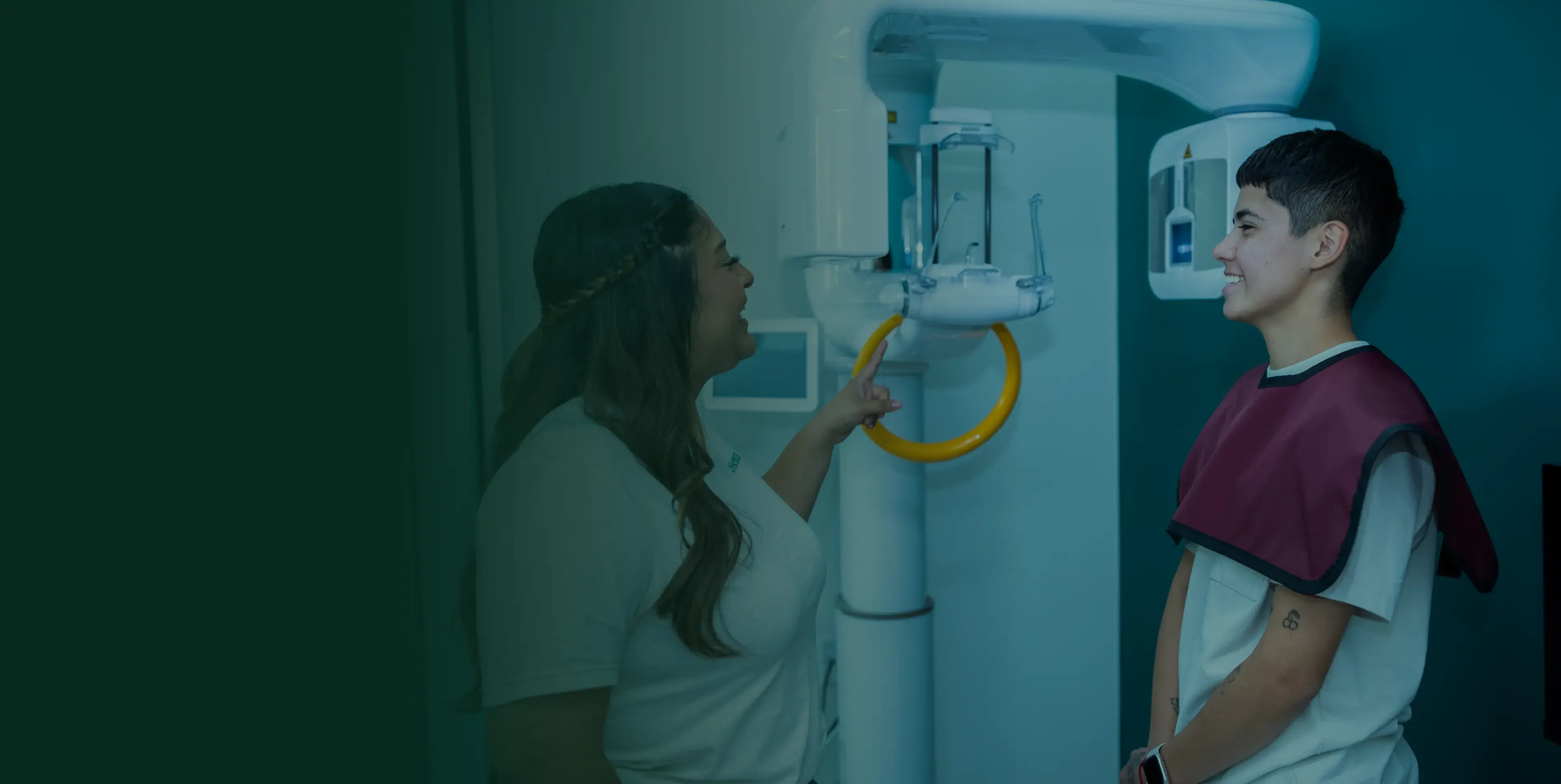 A woman points at a dental X-ray machine while a person stands nearby wearing a protective apron.