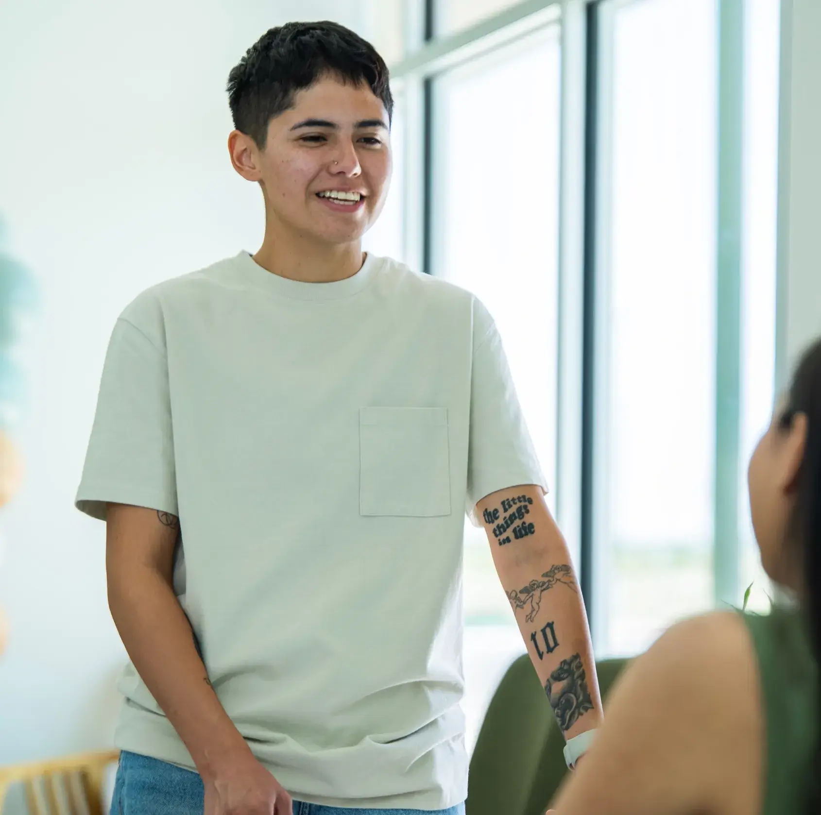 A person with tattoos on their arm smiles while talking to someone indoors.