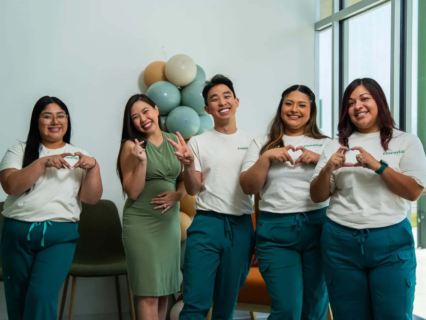 Five people smiling, making heart shapes with their hands, stand together in a bright room.