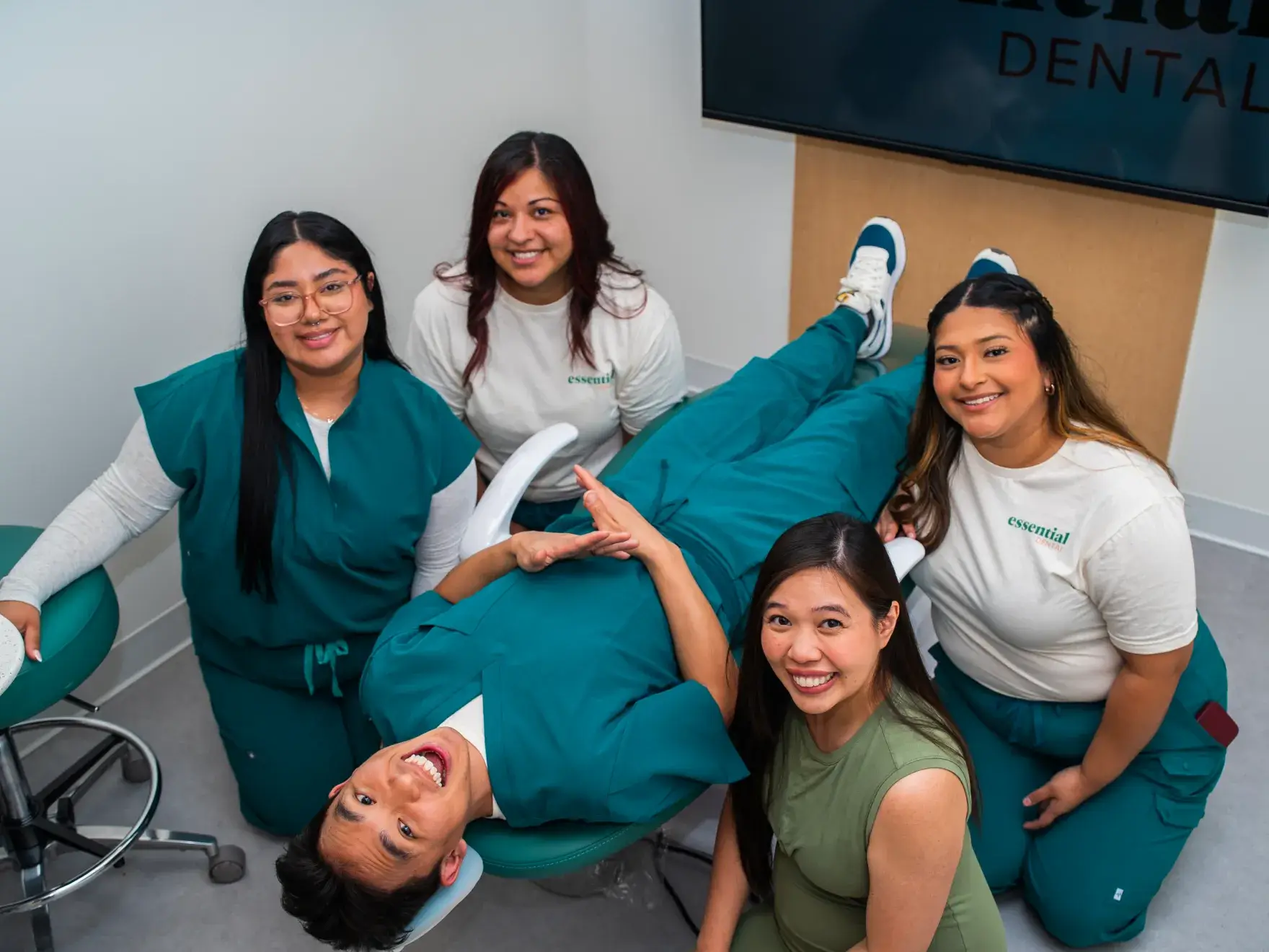 Five people smiling, one person lying on a dentist chair, in a dental office.