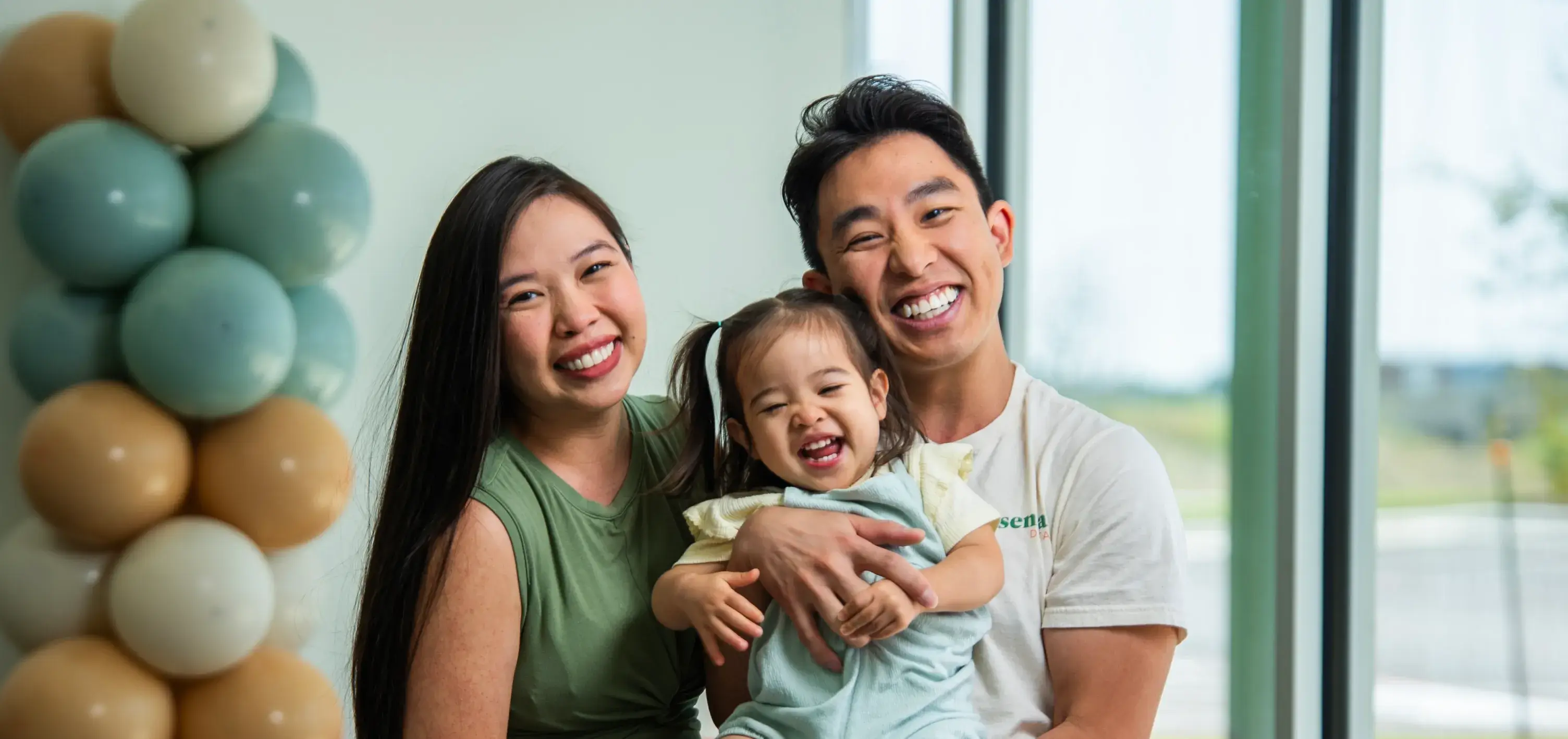 A smiling couple holds a happy toddler between them, next to a bunch of colorful balloons.