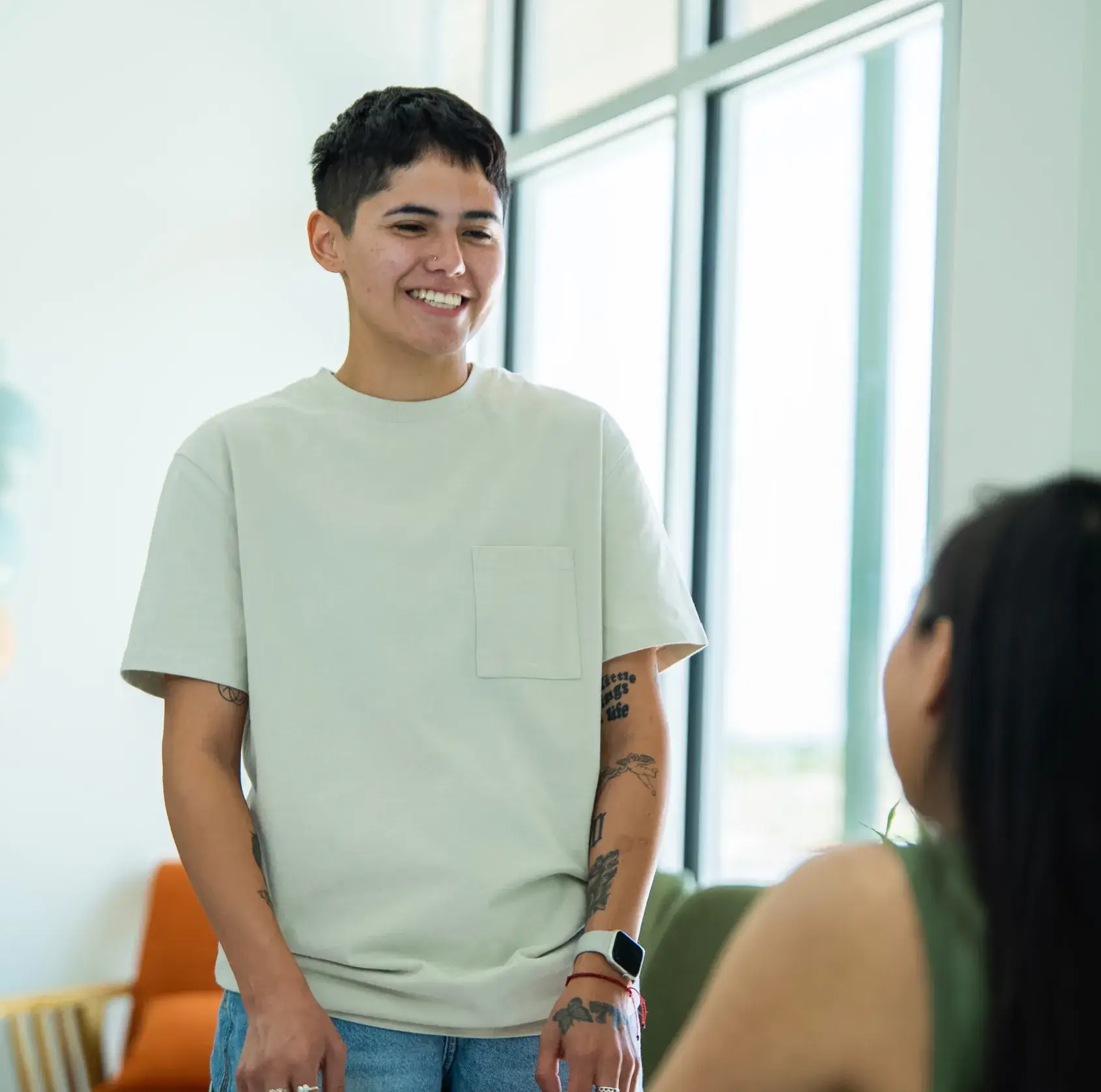 A person wearing a light shirt smiles and talks to a seated person.