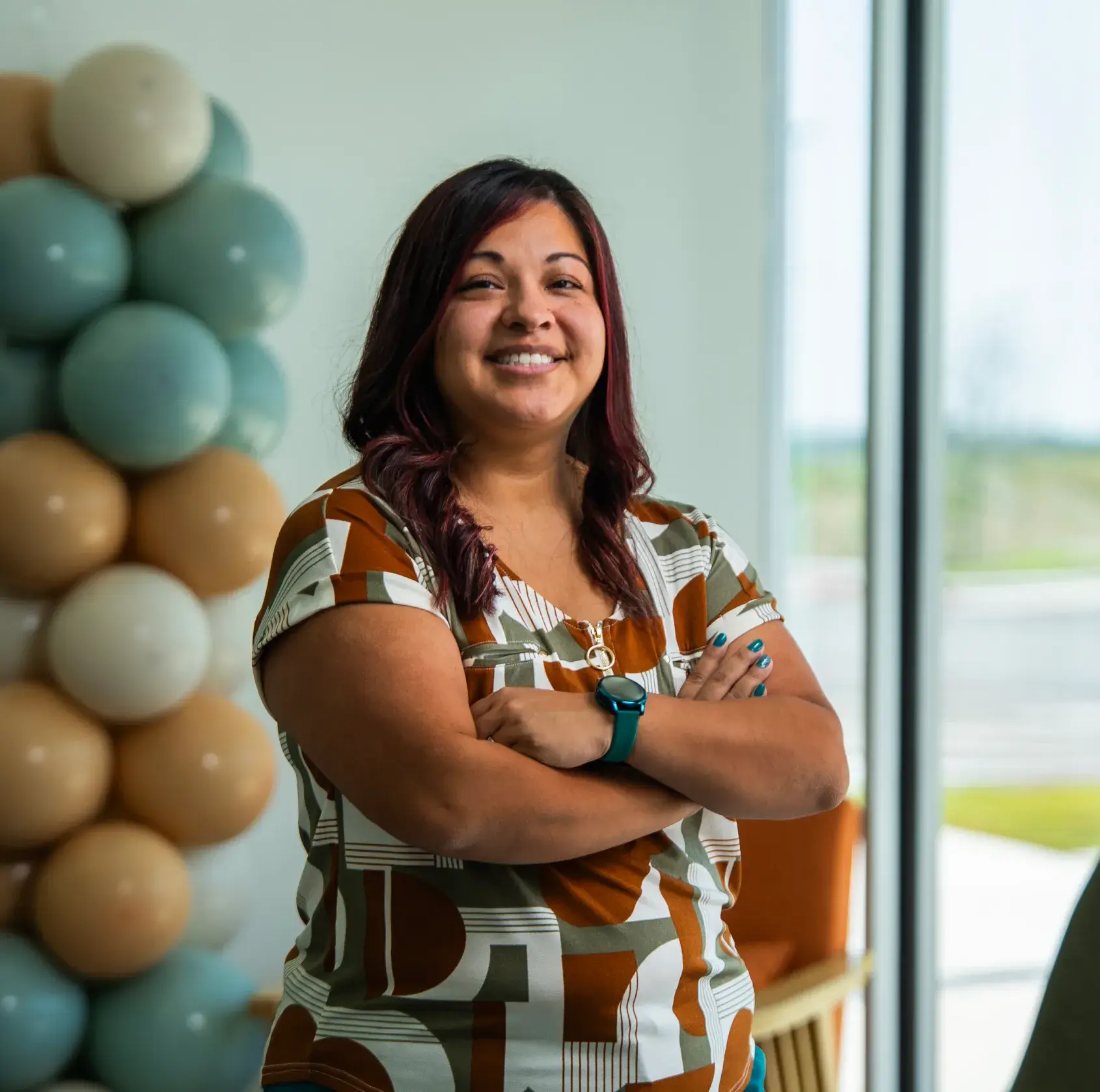 A woman with arms crossed is smiling indoors near a decorative balloon arrangement.